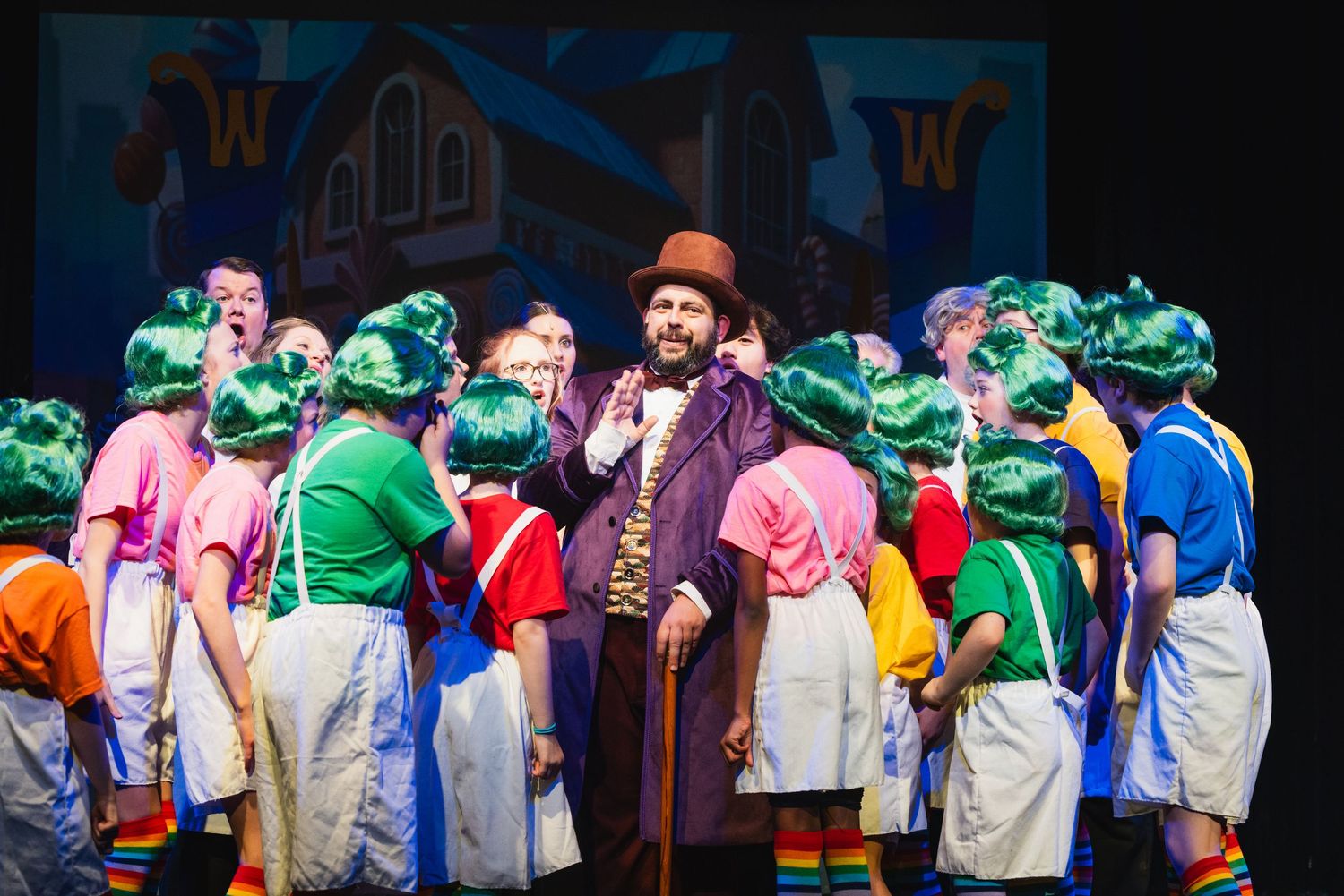 A Willy Wonka performer in a top hat poses with colorful Oompa Loompa characters on a theater stage.