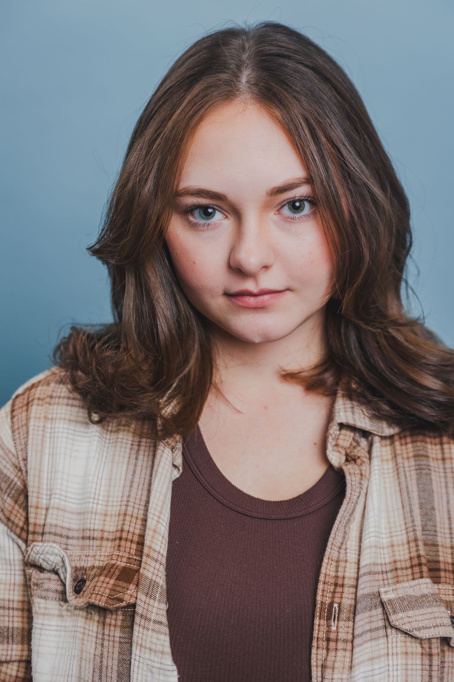 Young teenage girl with brown wavy hair wearing plaid shirt over brown top on gray background.