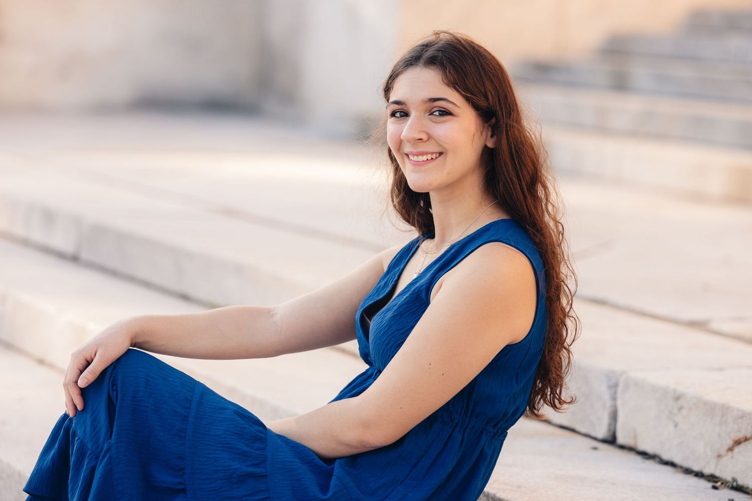 Young woman in blue dress sitting on stone steps, smiling outdoors.