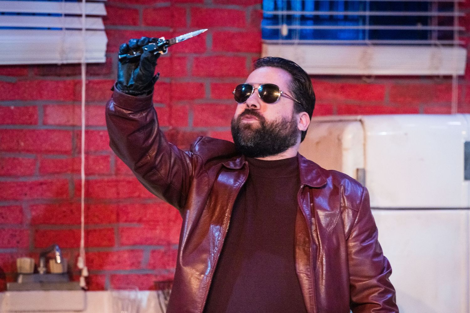 Bearded man in sunglasses and a brown leather jacket holding a knife dramatically in a theatrical performance scene.