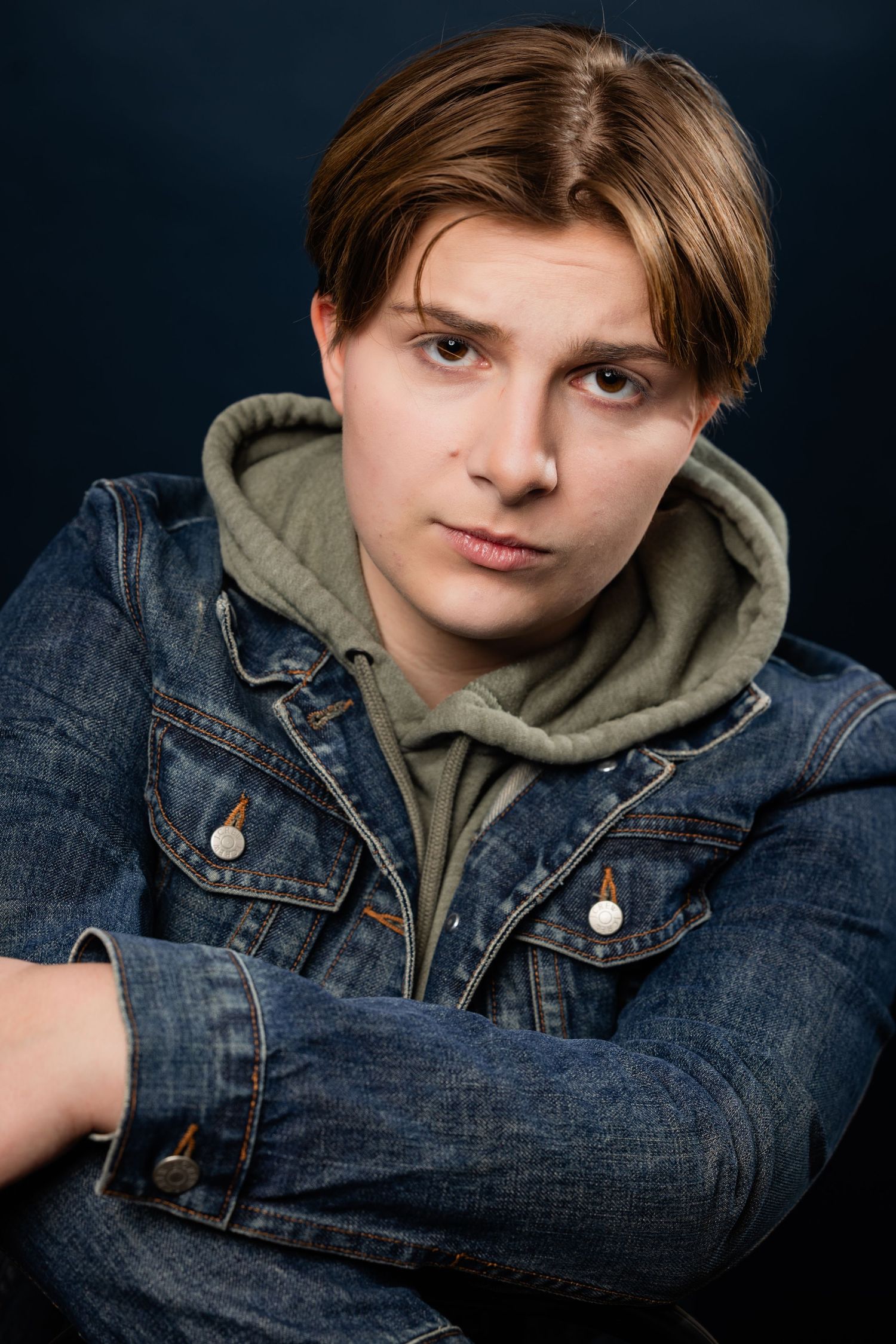 A young man with light brown hair wearing a denim jacket and hoodie poses against a dark background.