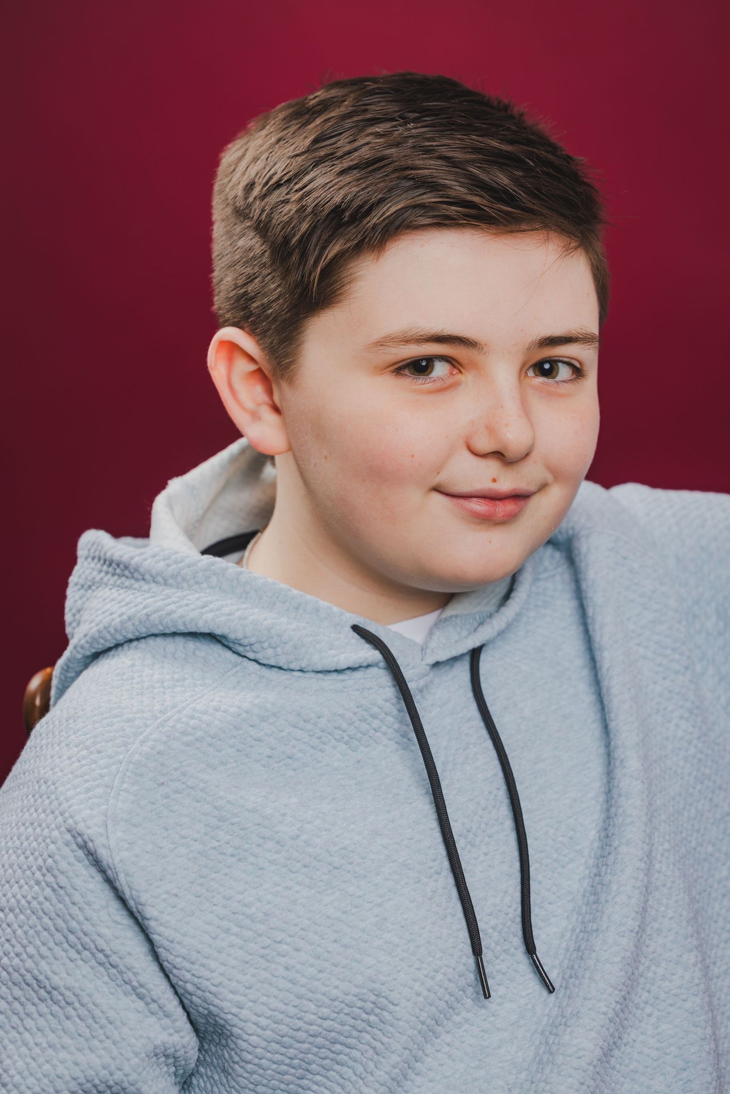Smiling preteen boy with short brown hair wearing a light blue hoodie against a red background.