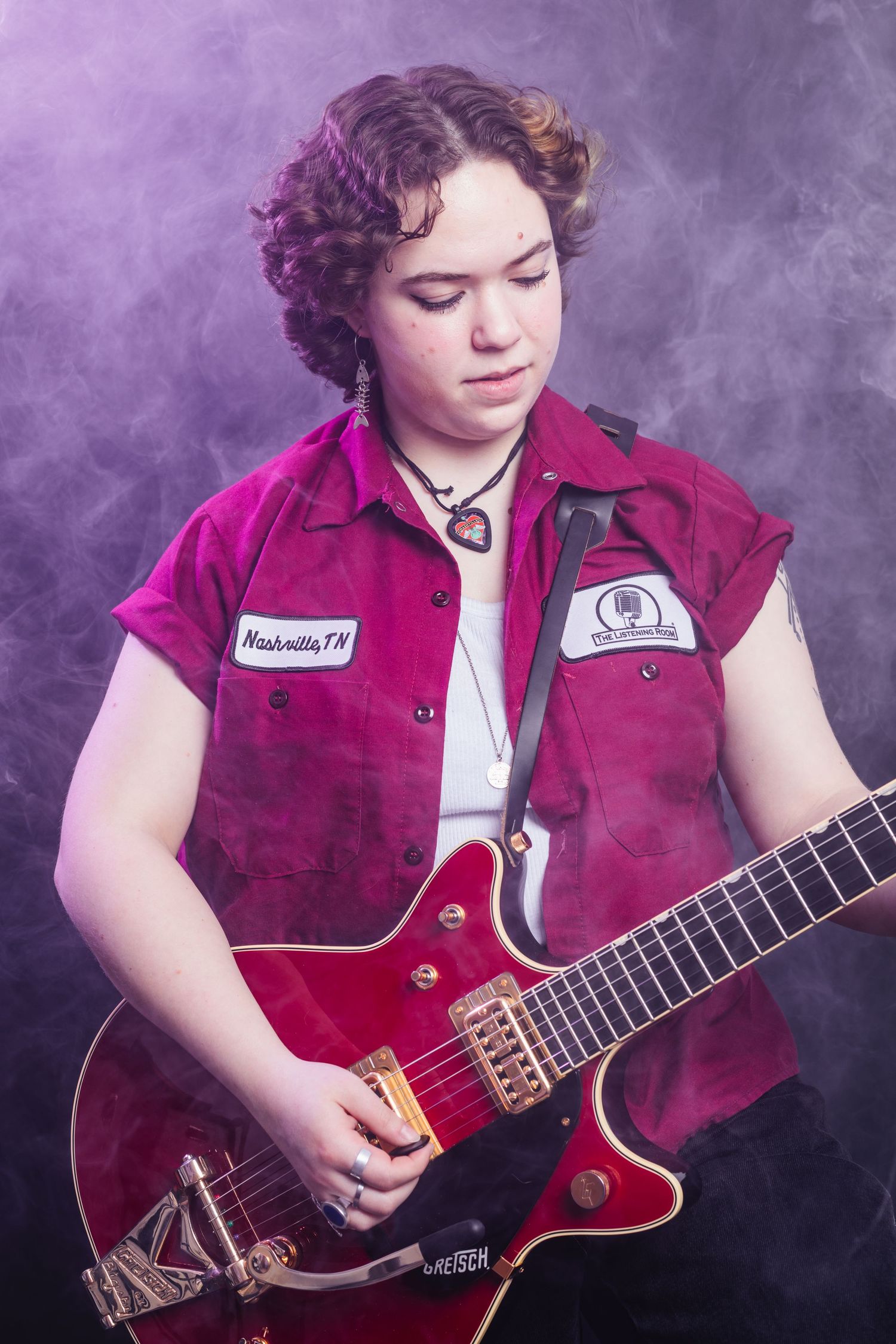 Young musician playing a red electric guitar in a purple smoky studio, wearing a Nashville TN patch shirt.