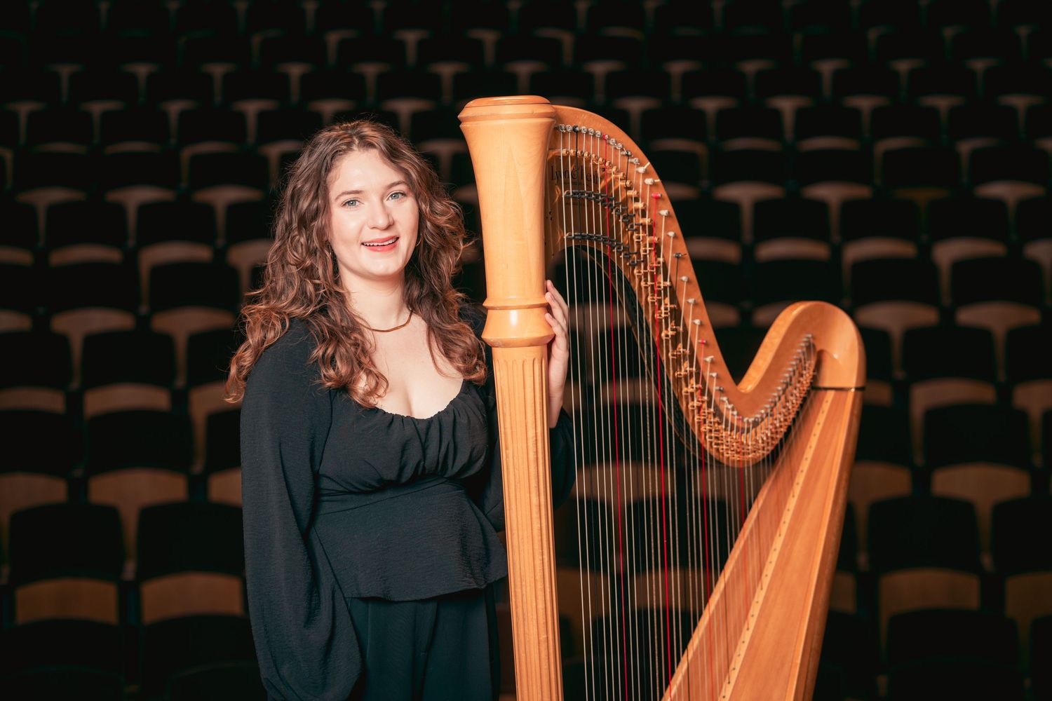 Young woman with curly hair posing beside a concert harp in a performance hall.