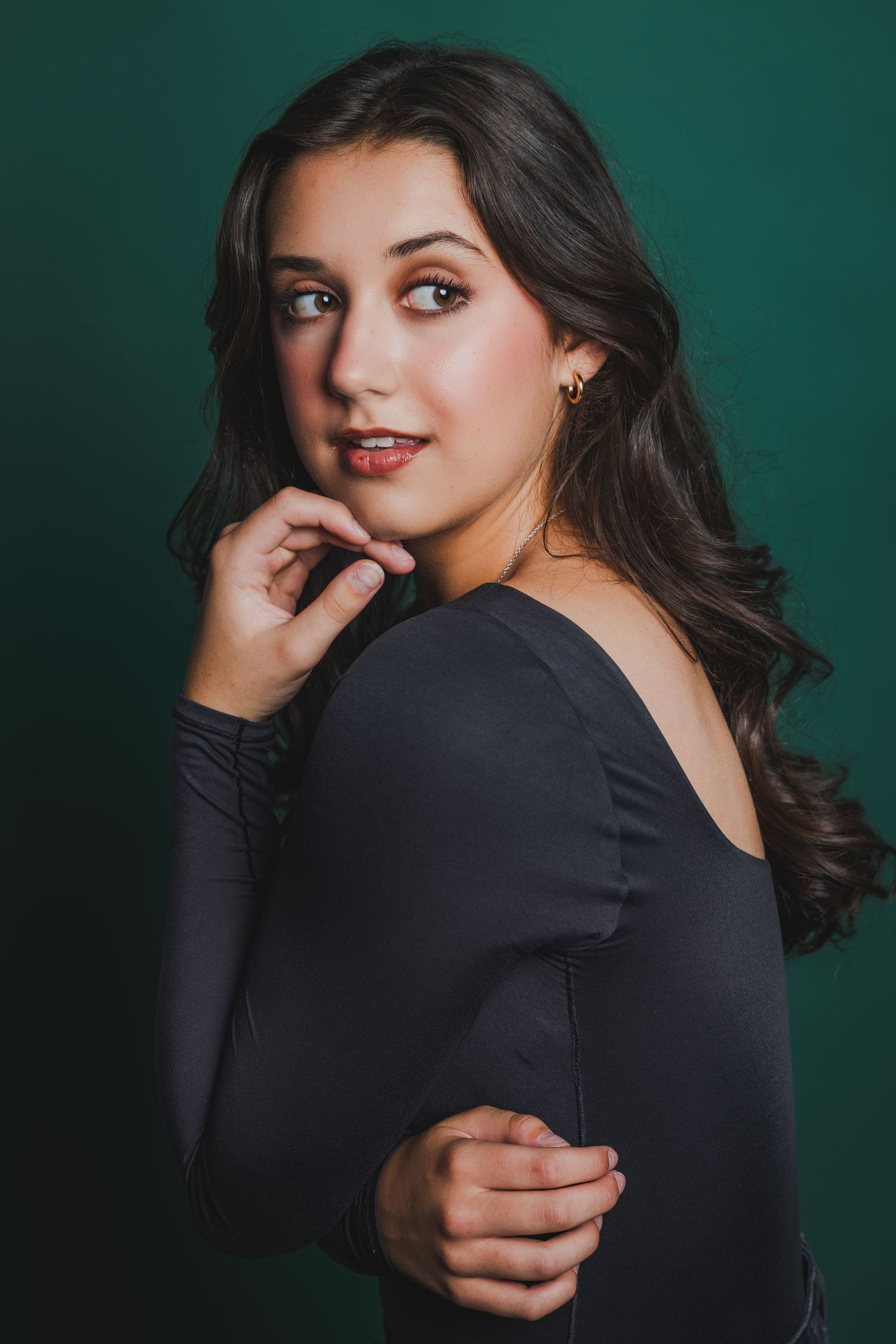 Young brunette woman in black off-shoulder top posing against teal studio background.