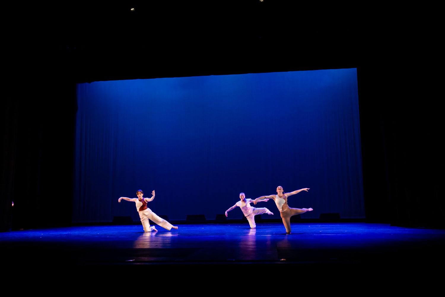 Three ballet dancers perform an elegant routine under dramatic blue stage lighting in a dark theater.