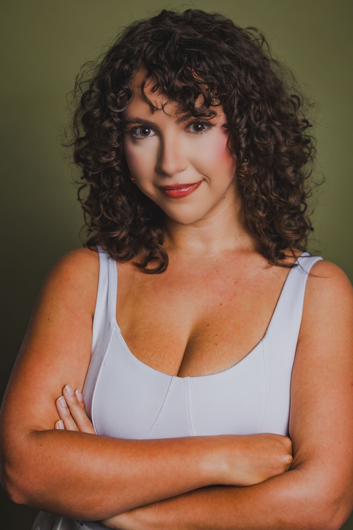 Woman with curly brown hair wearing white tank top with arms crossed on green background.