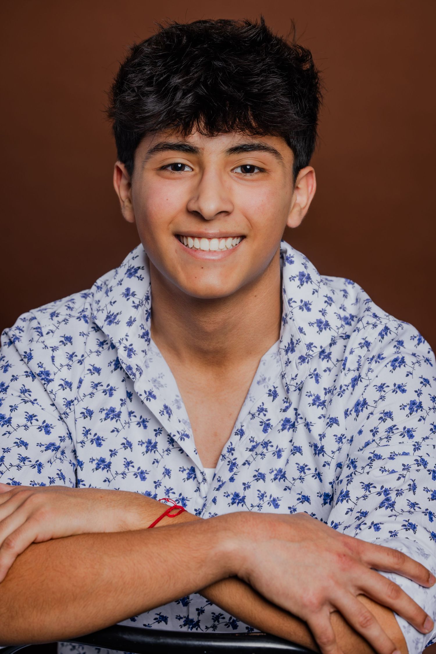 Smiling teenage boy with dark curly hair wearing a floral print shirt, arms crossed on brown background.