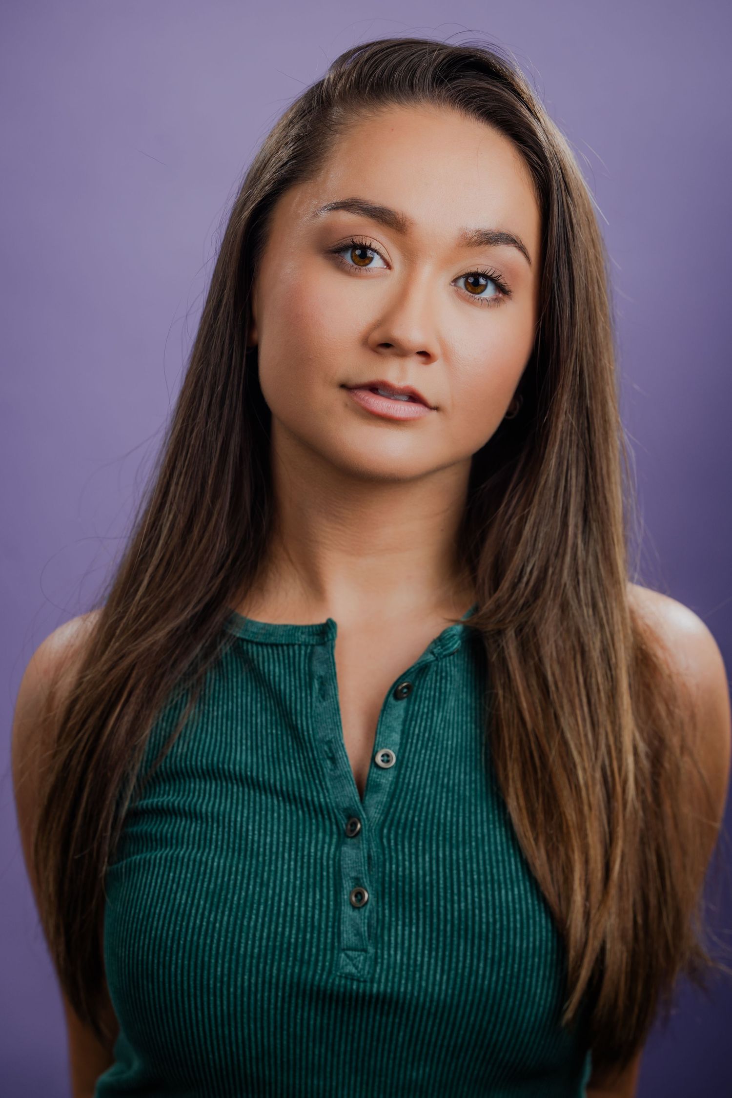 Young woman with long straight brown hair wearing a green ribbed henley top against a purple background.