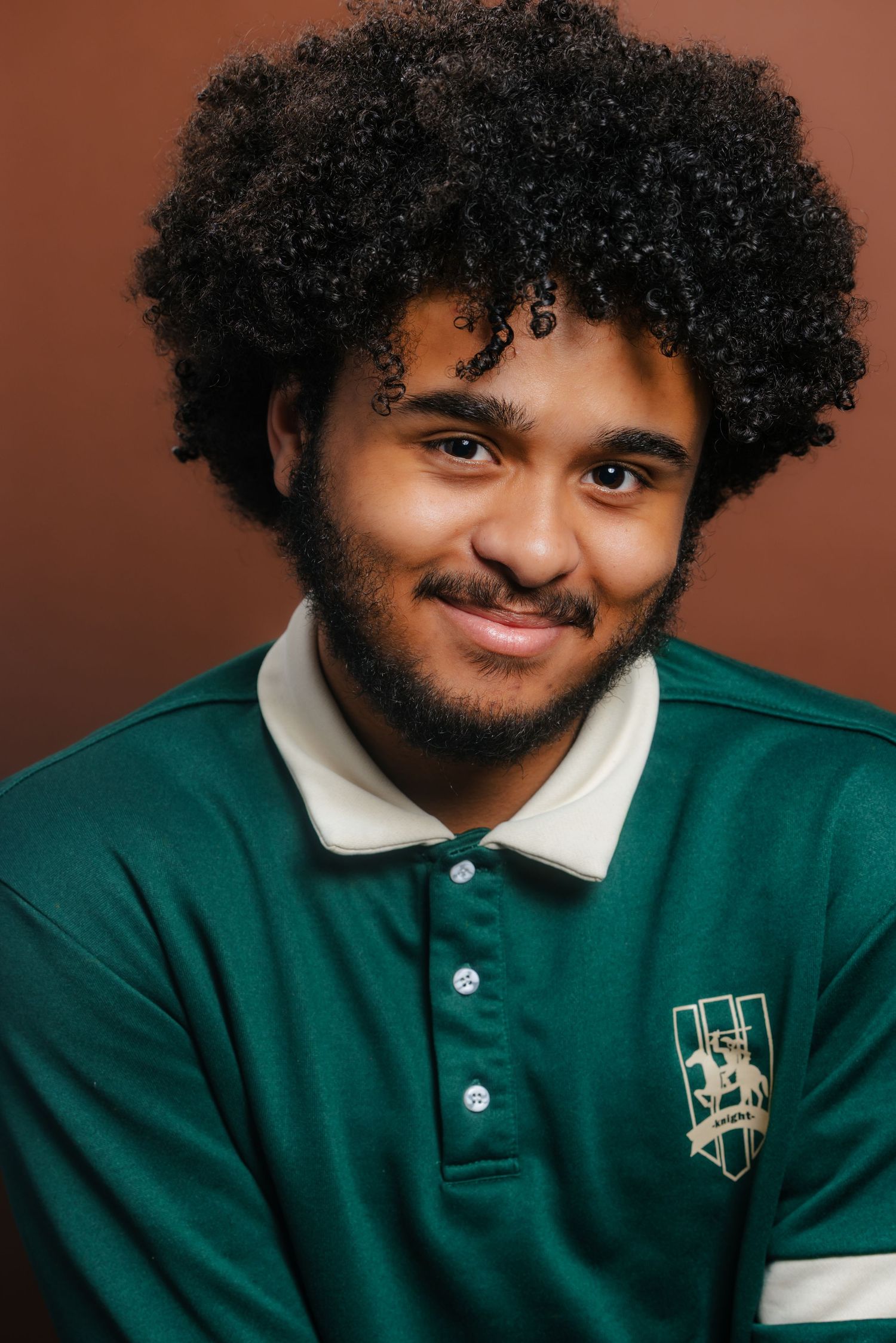 Smiling young man with a curly afro wearing a green polo shirt against a warm brown background.