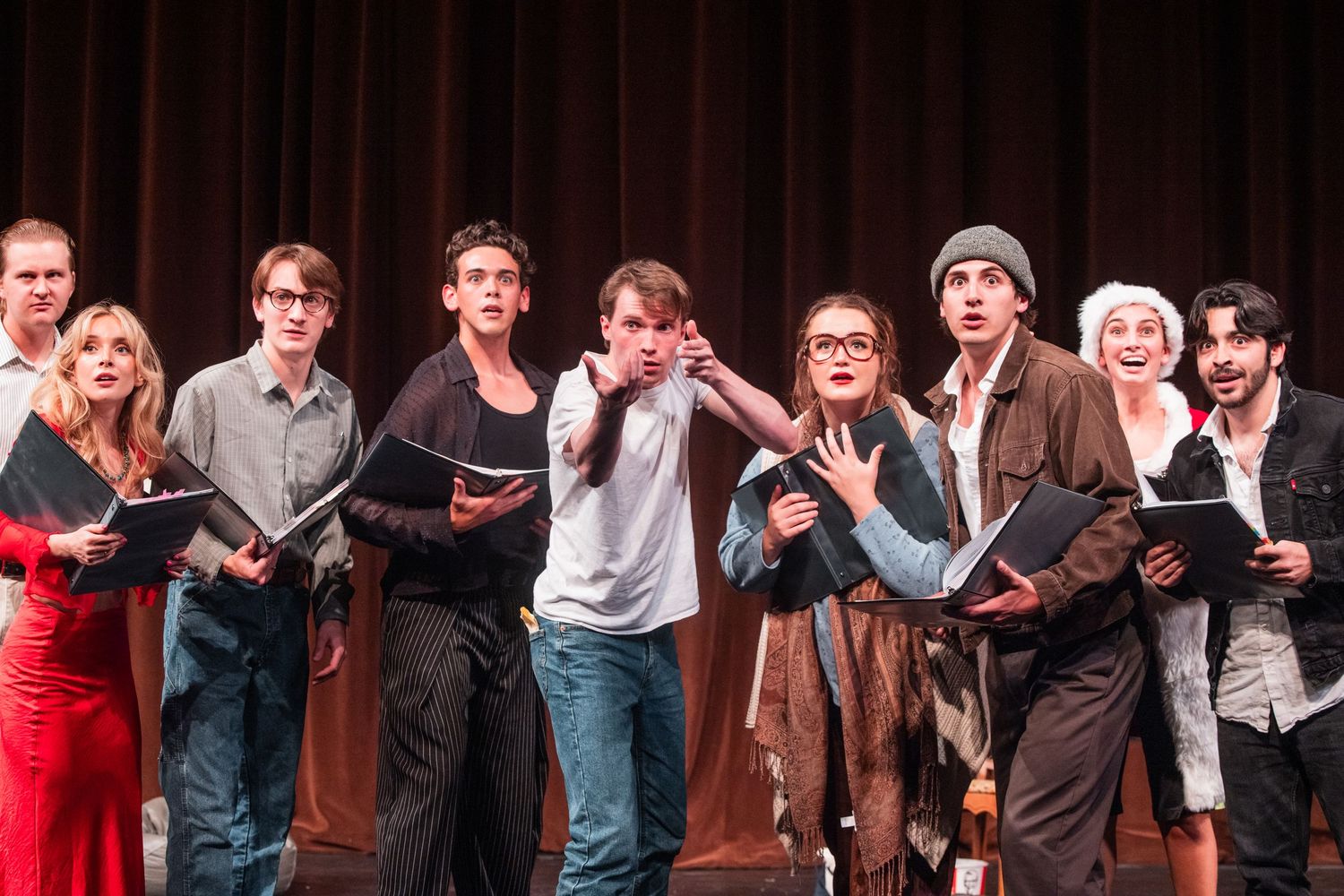 Large theater ensemble cast performing dramatically on stage holding black scripts under stage lights.
