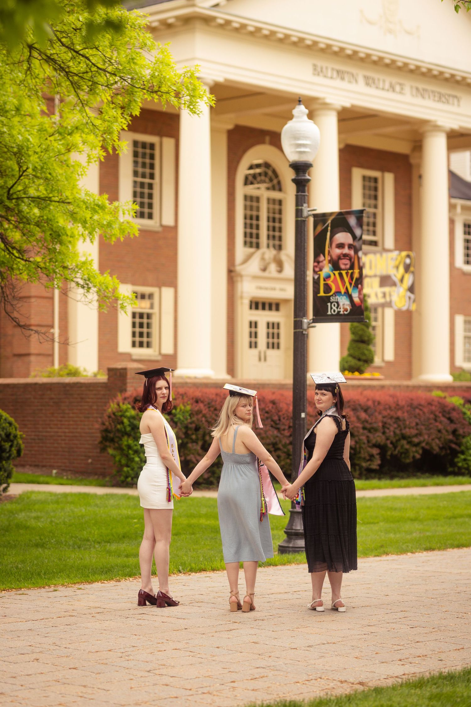 Three female graduates holding hands on campus path at Baldwin Wallace University.