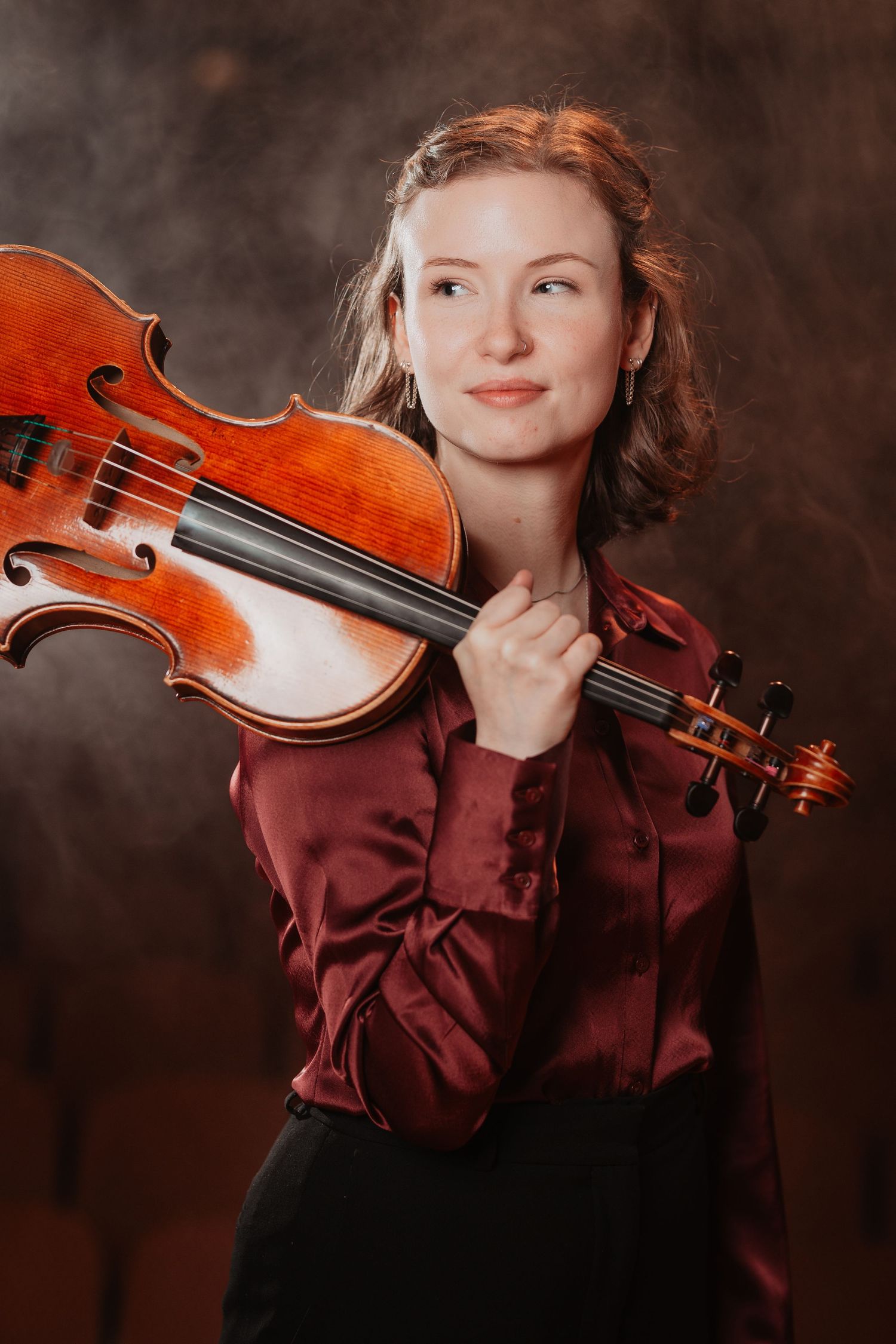 Young female violinist in a burgundy silk shirt holding a violin on her shoulder against a moody dark background.