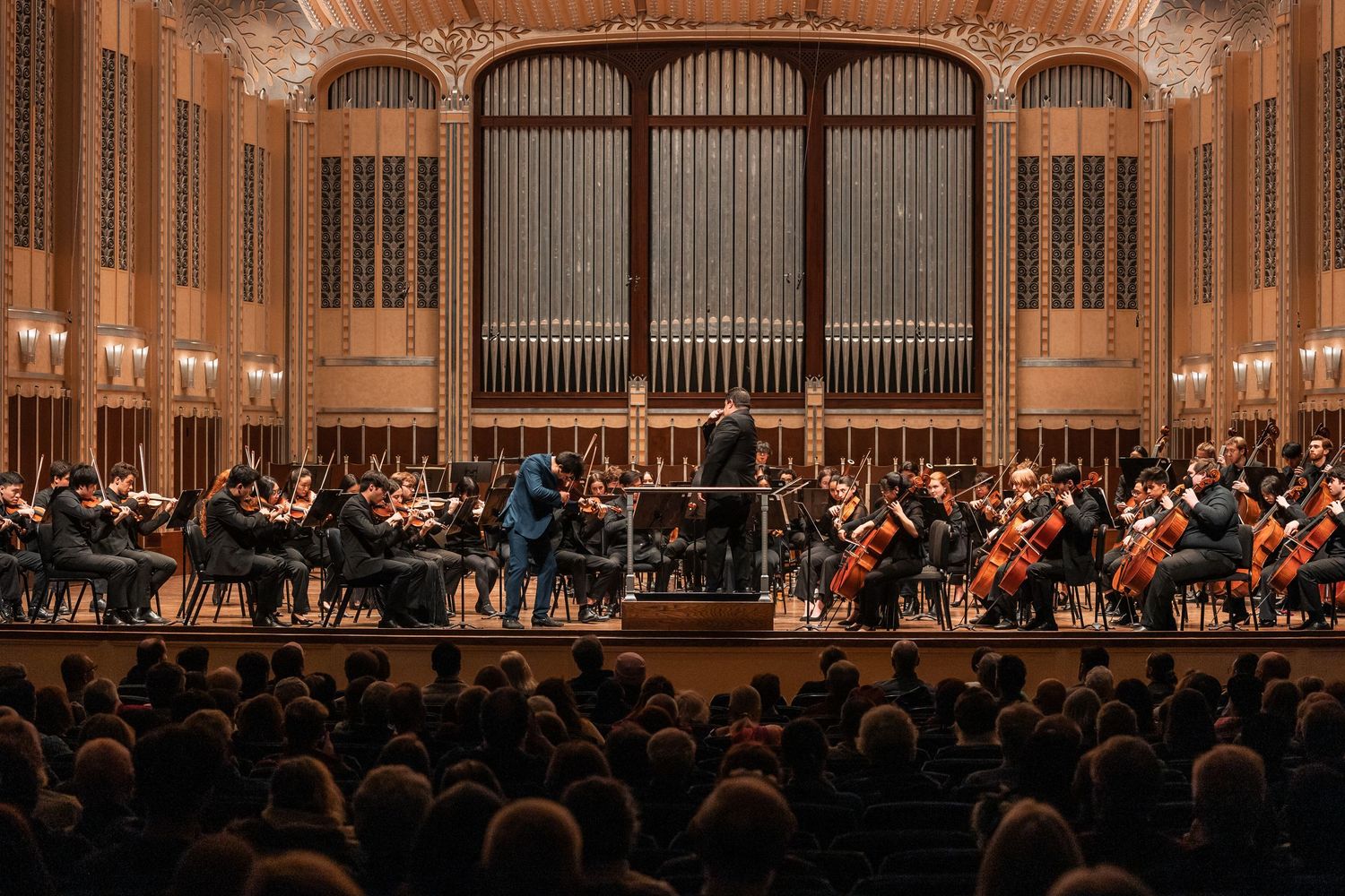 A full orchestra performs on stage in an ornate concert hall with a conductor leading the ensemble.