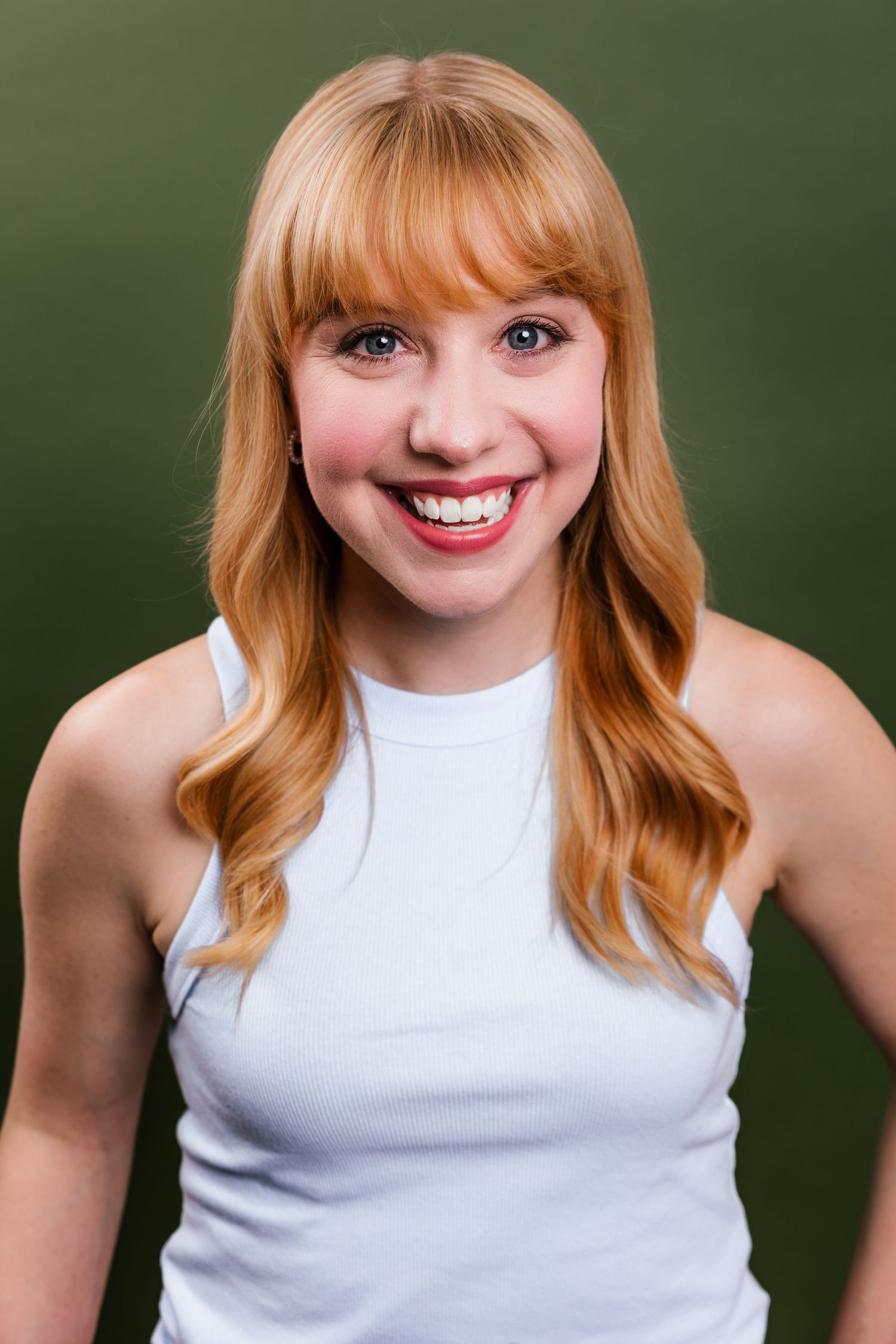 Cheerful young woman with long blonde hair smiles brightly in a white tank top on a green background.