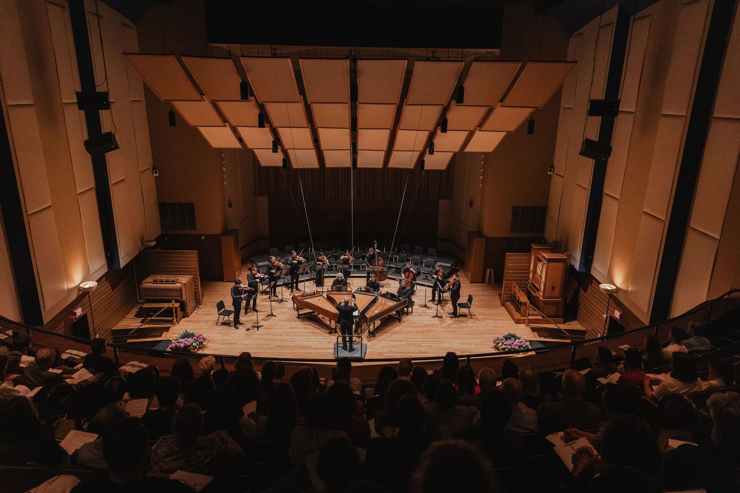 Aerial view of a full orchestra performing on stage in a modern concert hall with audience seated below.