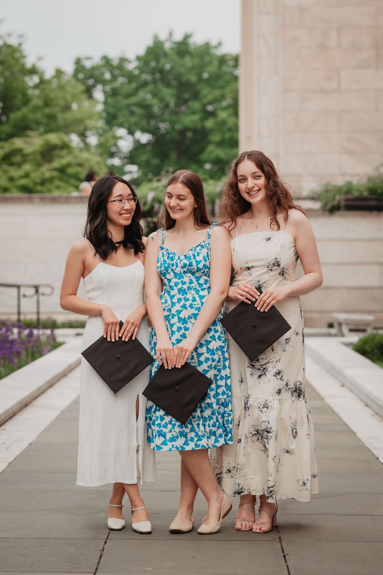 Three female graduates in summer dresses holding mortarboard caps in a garden setting.