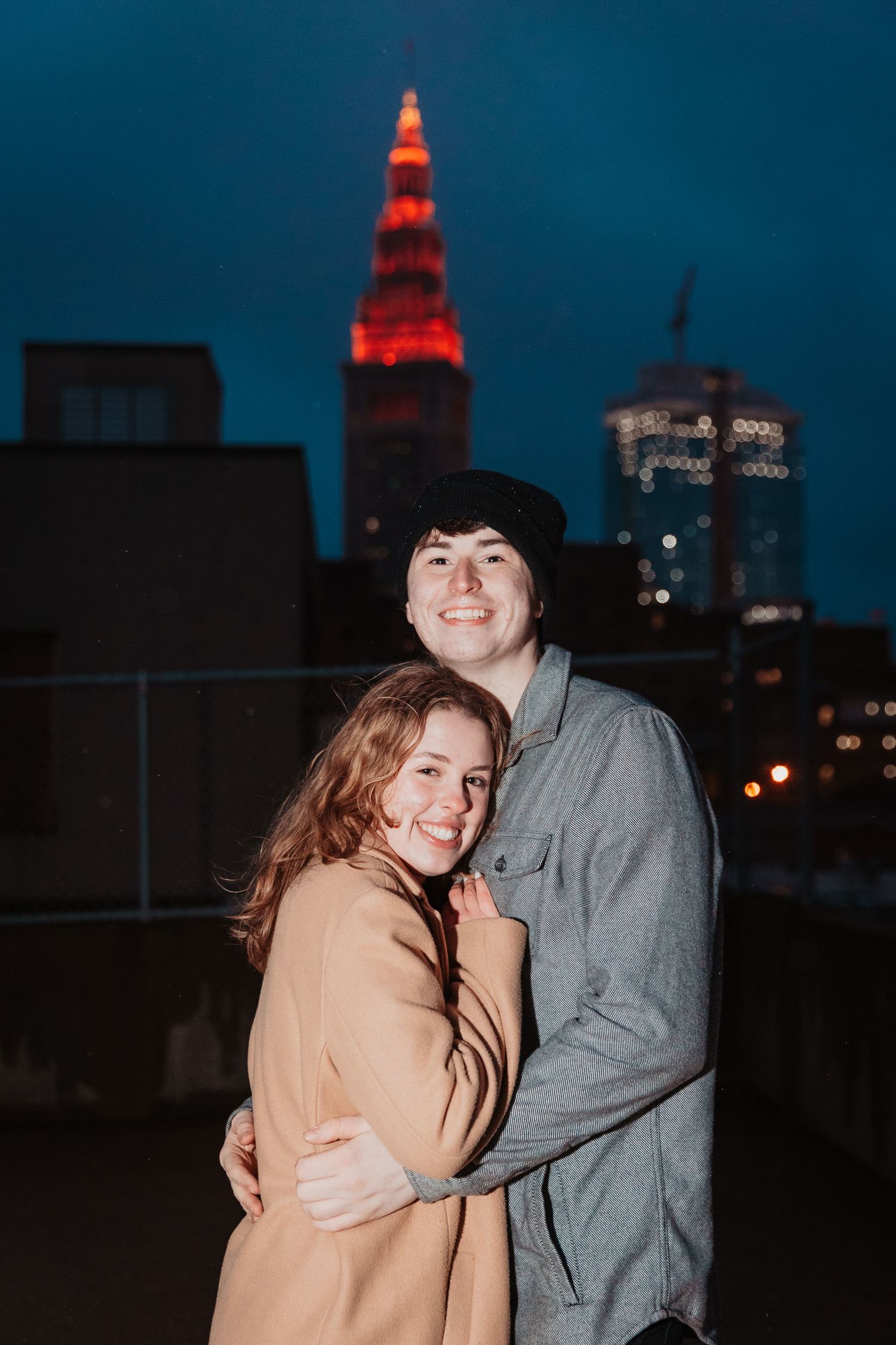 Smiling couple embracing on rooftop at night with illuminated red Terminal Tower in background.