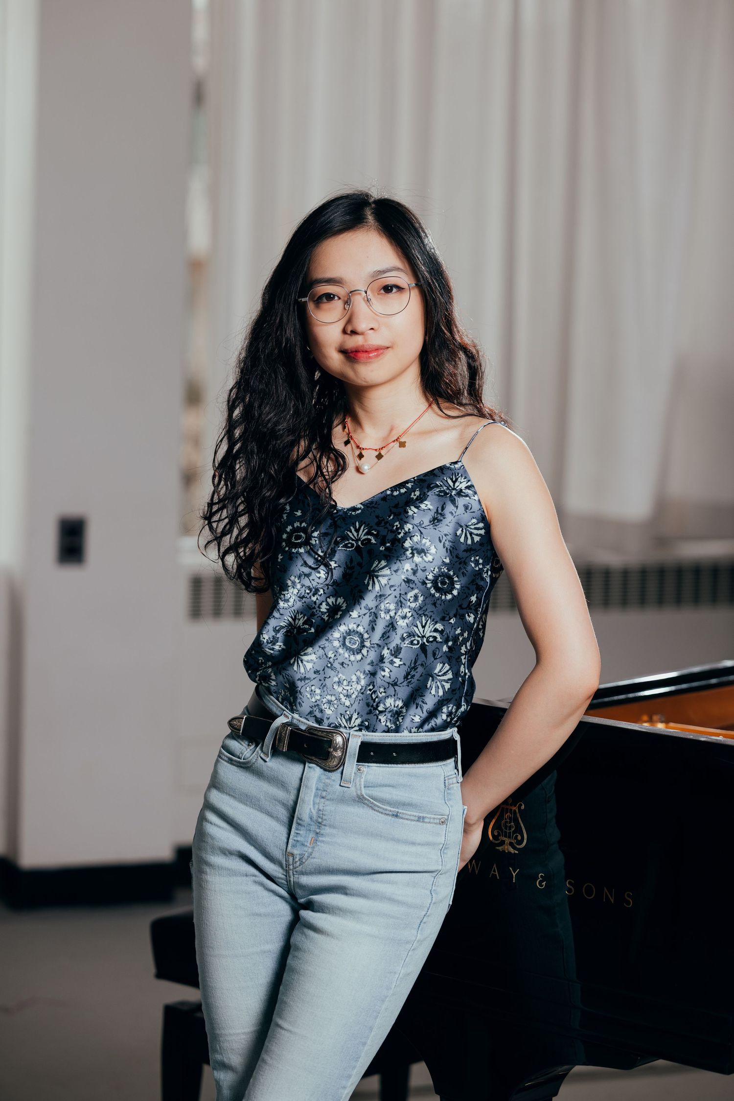 Young Asian female pianist with glasses leaning against a grand piano in a bright airy studio space.