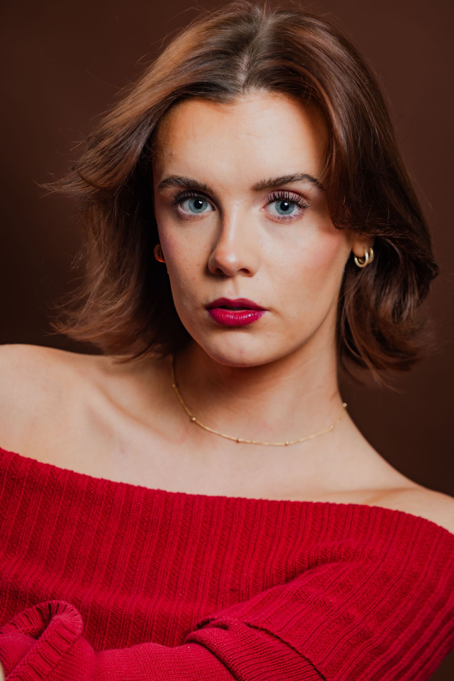 Young woman with short brown hair and blue eyes wearing a red off-shoulder sweater against dark background.