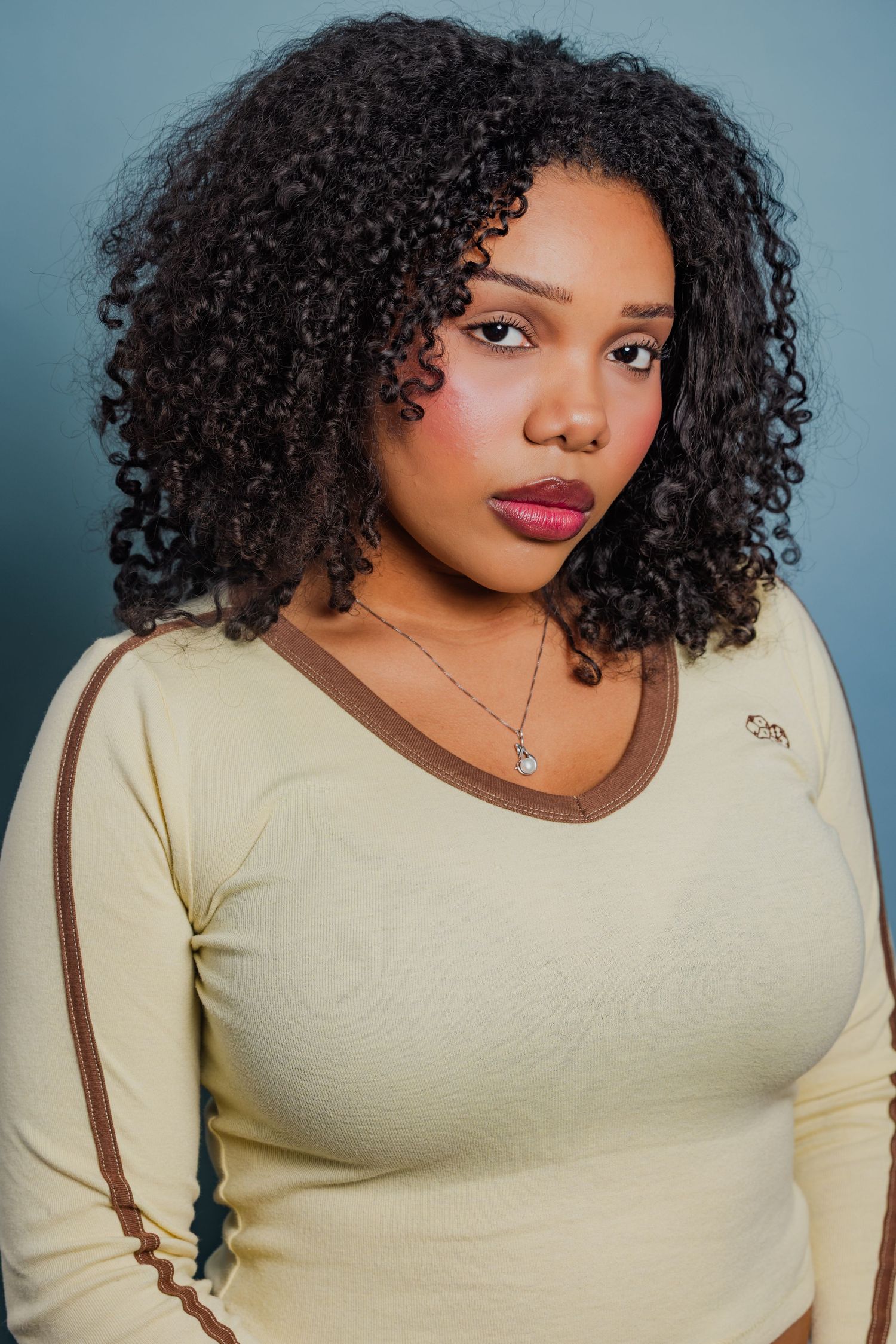 Young woman with curly black hair wearing cream long-sleeve top and necklace, studio portrait.
