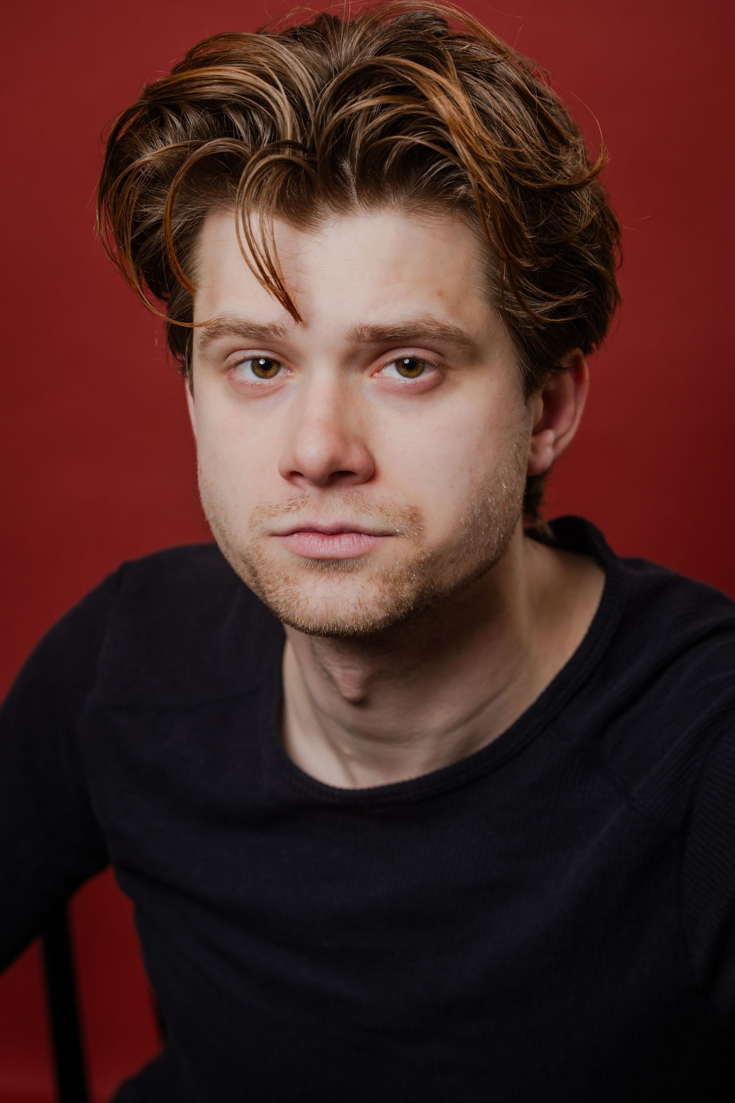 A young man with highlighted brown hair wearing a black t-shirt poses against a red background.