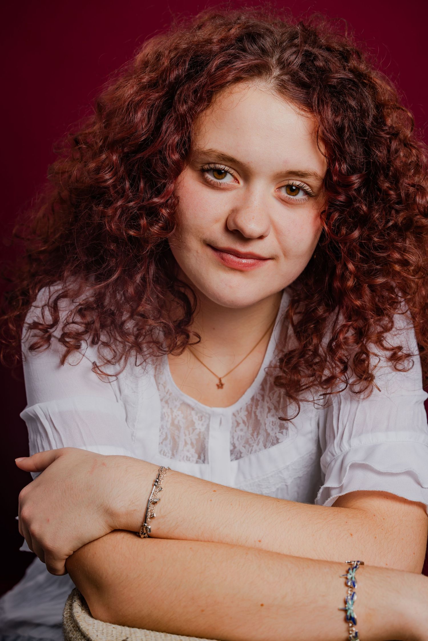 Young woman with curly red hair wearing white blouse and bracelets, studio portrait photo.