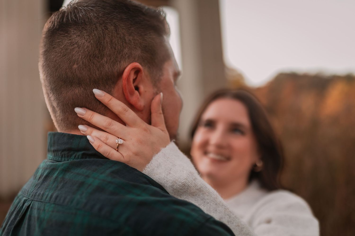 Close-up of a smiling woman touching a man's face, showcasing an engagement ring during a fall photo session.