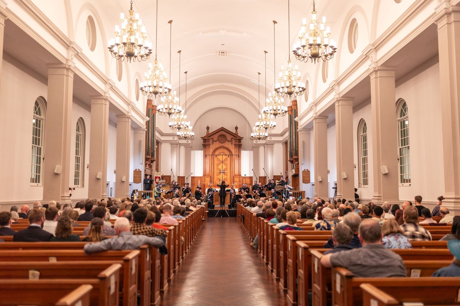 An orchestra performs for a seated audience inside an elegant white church with chandeliers.
