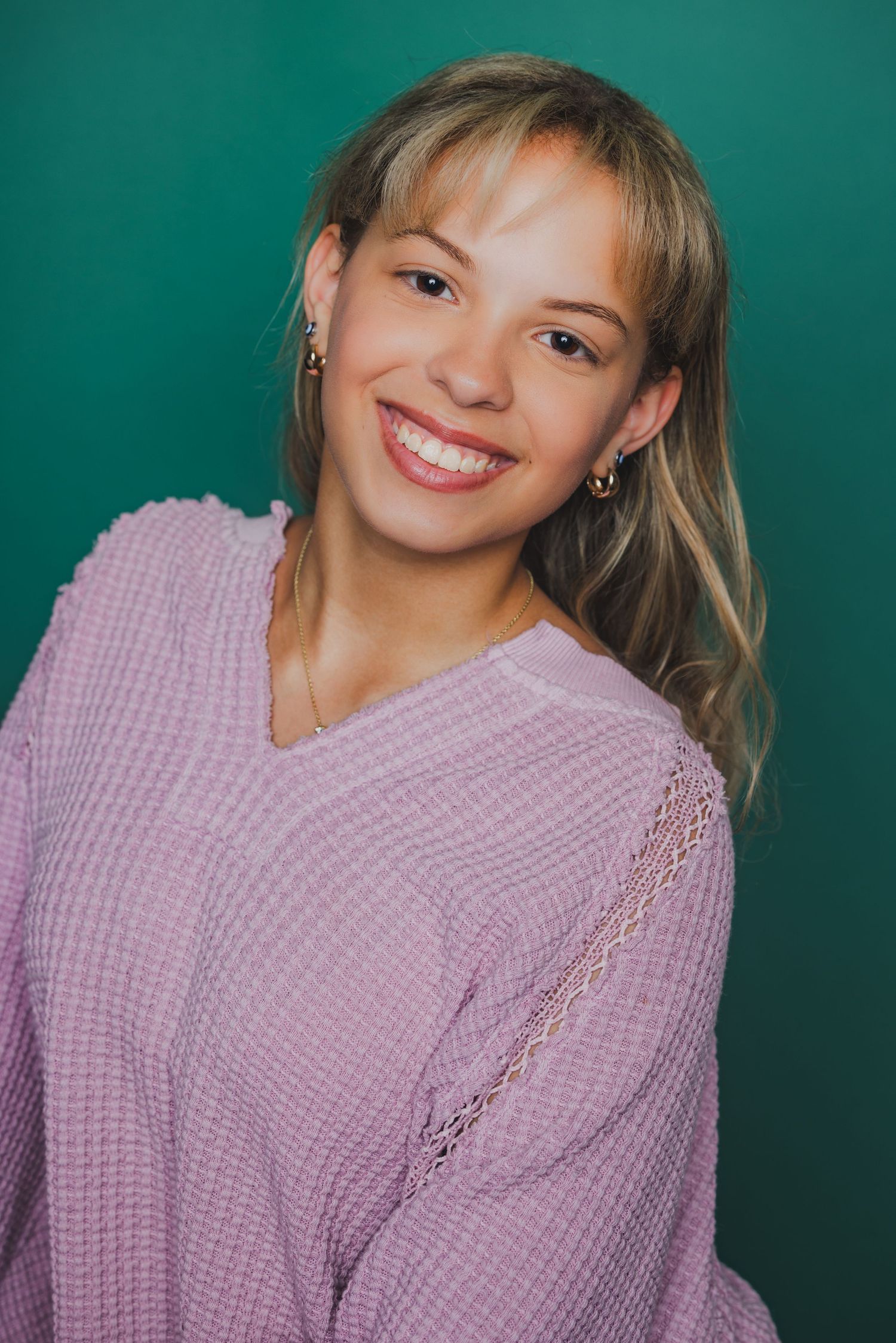 Teenage girl with blonde hair smiling in a lavender sweater against a teal background.