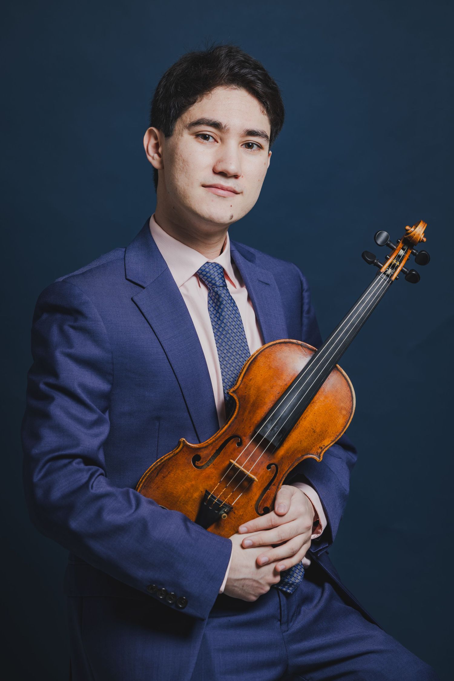 Young male violinist in a blue suit holding his violin, posing for a portrait on a dark blue background.