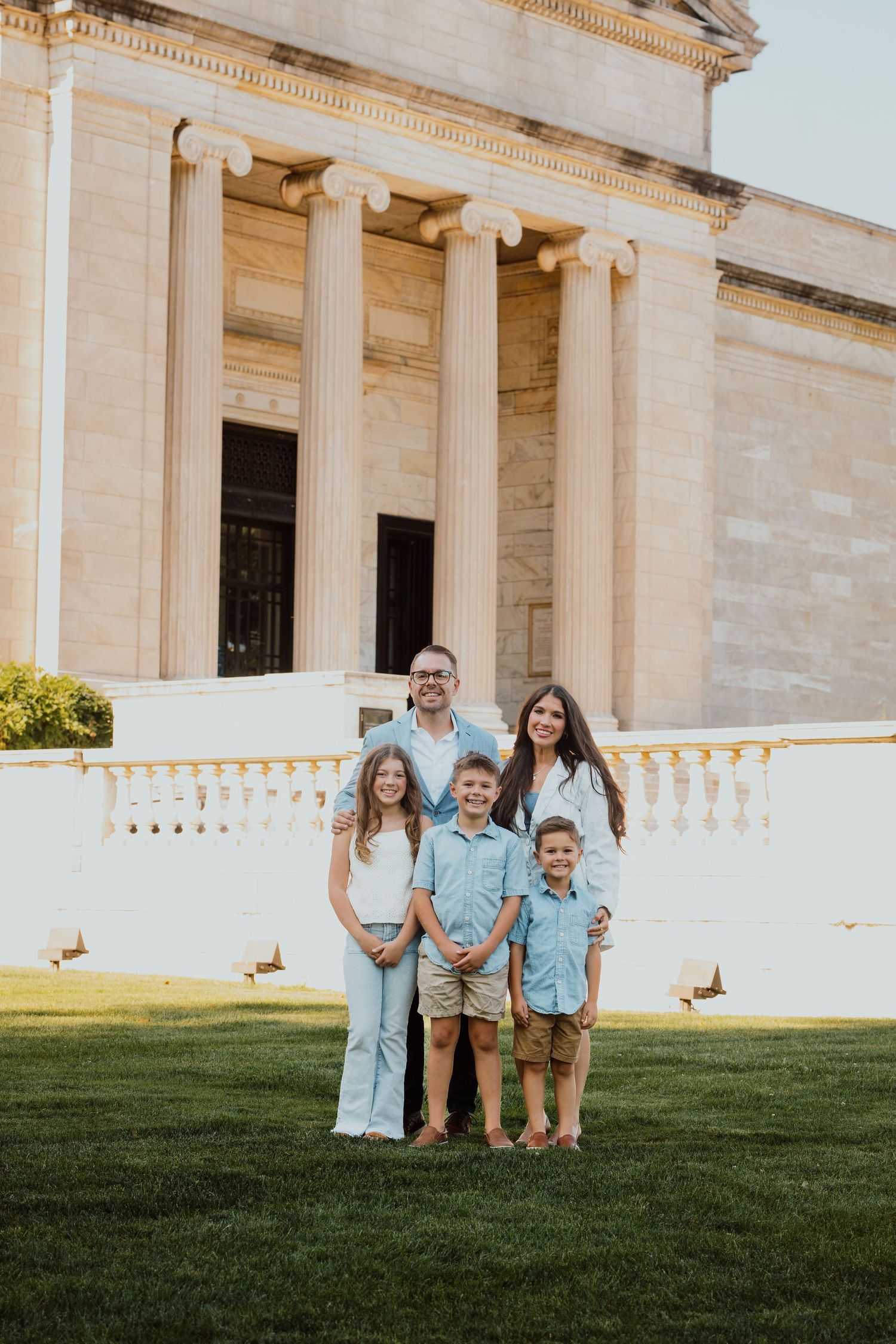 A happy family of five poses on green grass before a grand neoclassical building.