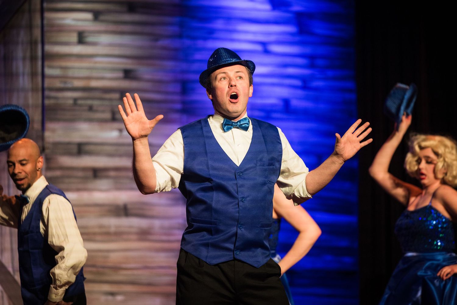 A male performer in a blue vest and bow tie gestures expressively on a theatrical stage.