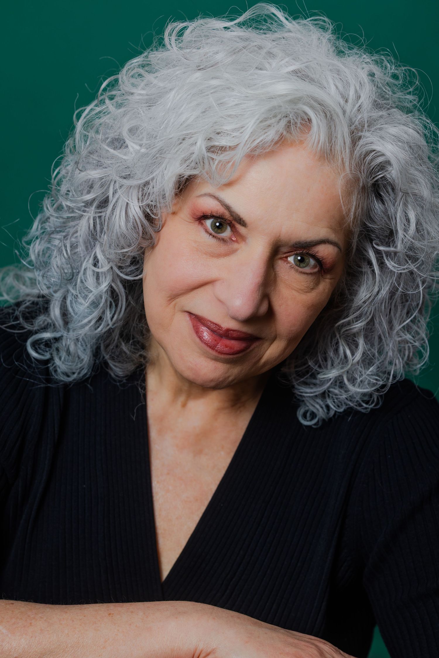 An older woman with curly gray hair wearing a black v-neck top poses against a green background.