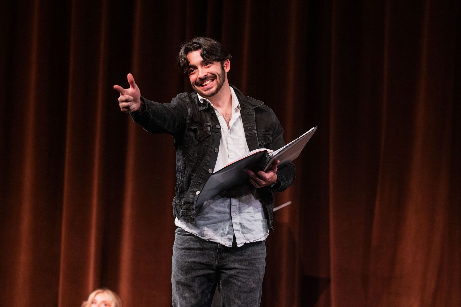 Smiling male actor holding a script and pointing at audience on a theater stage.