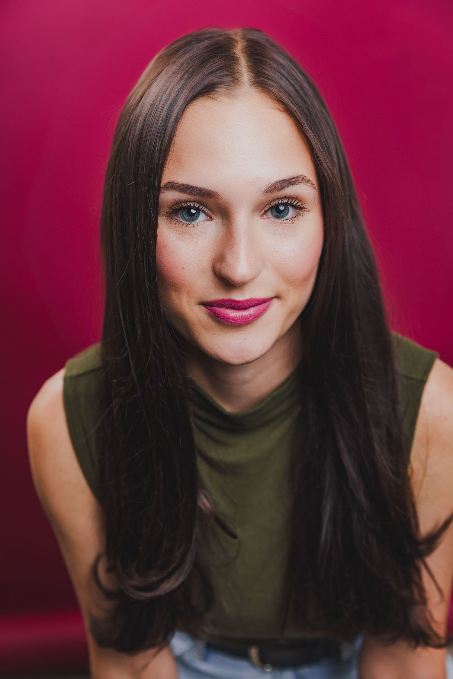 Young woman with long dark hair and blue eyes smiles in a green top against a pink background.