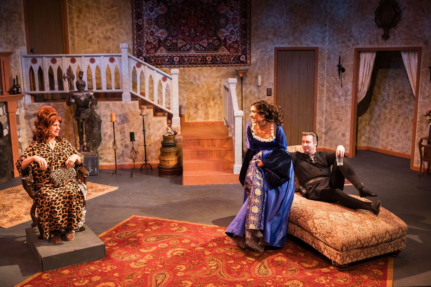 Three actors perform in an ornate living room stage set with a staircase and decorative rugs.