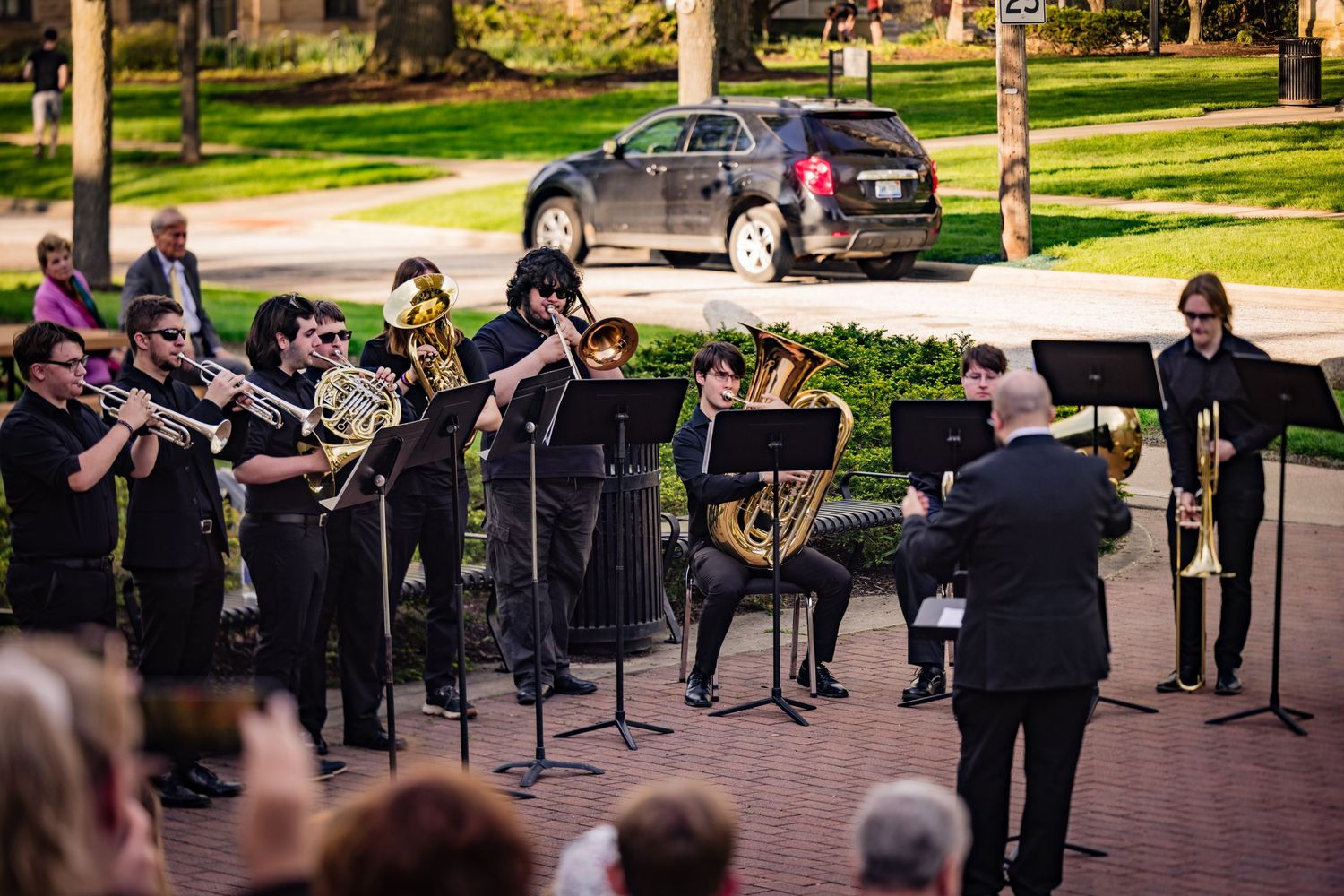 Brass ensemble performing outdoors on a campus walkway with a conductor leading them.