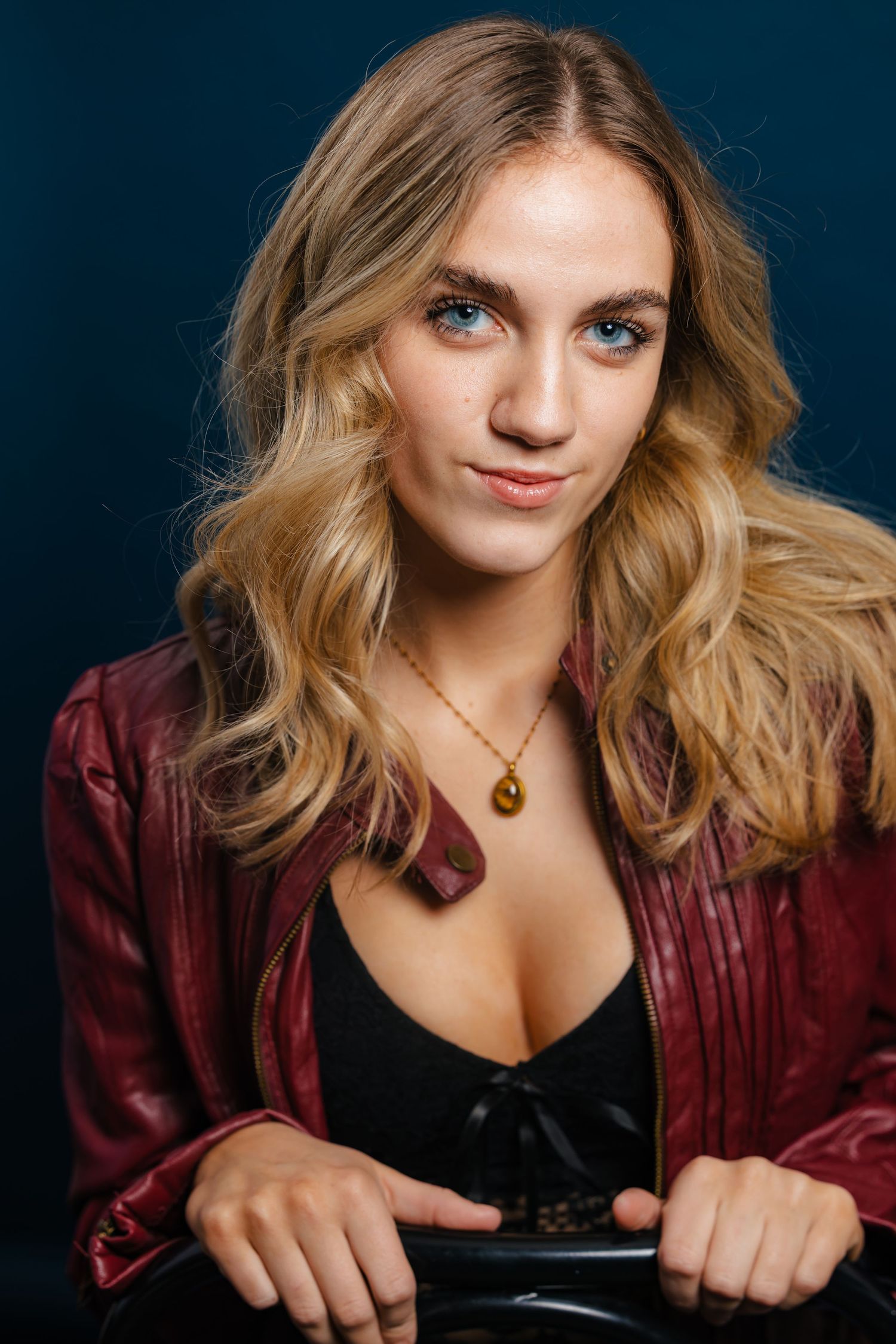 Young blonde woman with blue eyes wearing a red leather jacket and gold necklace against dark background.