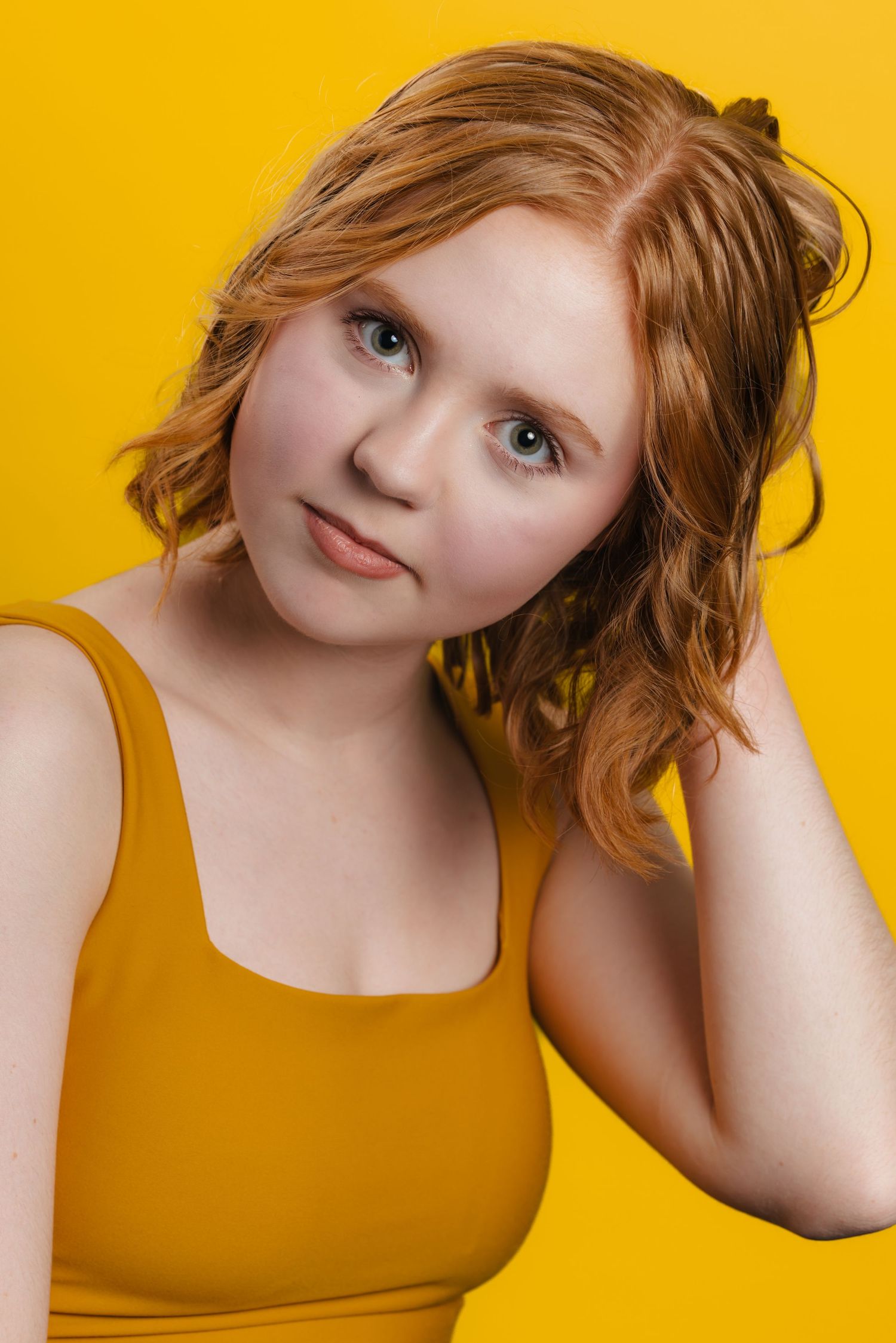Young woman with wavy red hair wearing a mustard yellow top posed against a matching yellow background.