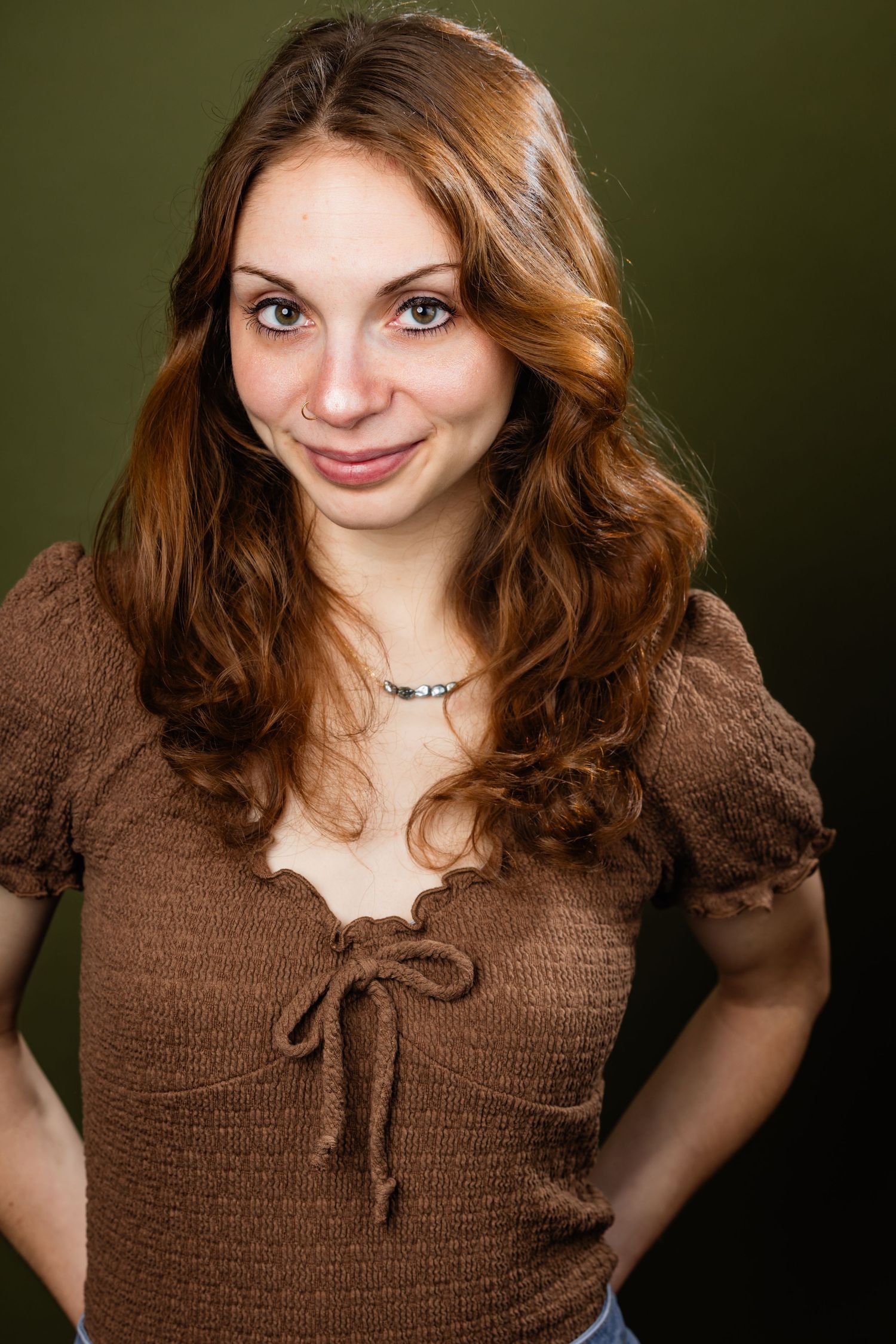 Young woman with auburn wavy hair smiles warmly wearing a brown knit top against dark green background.
