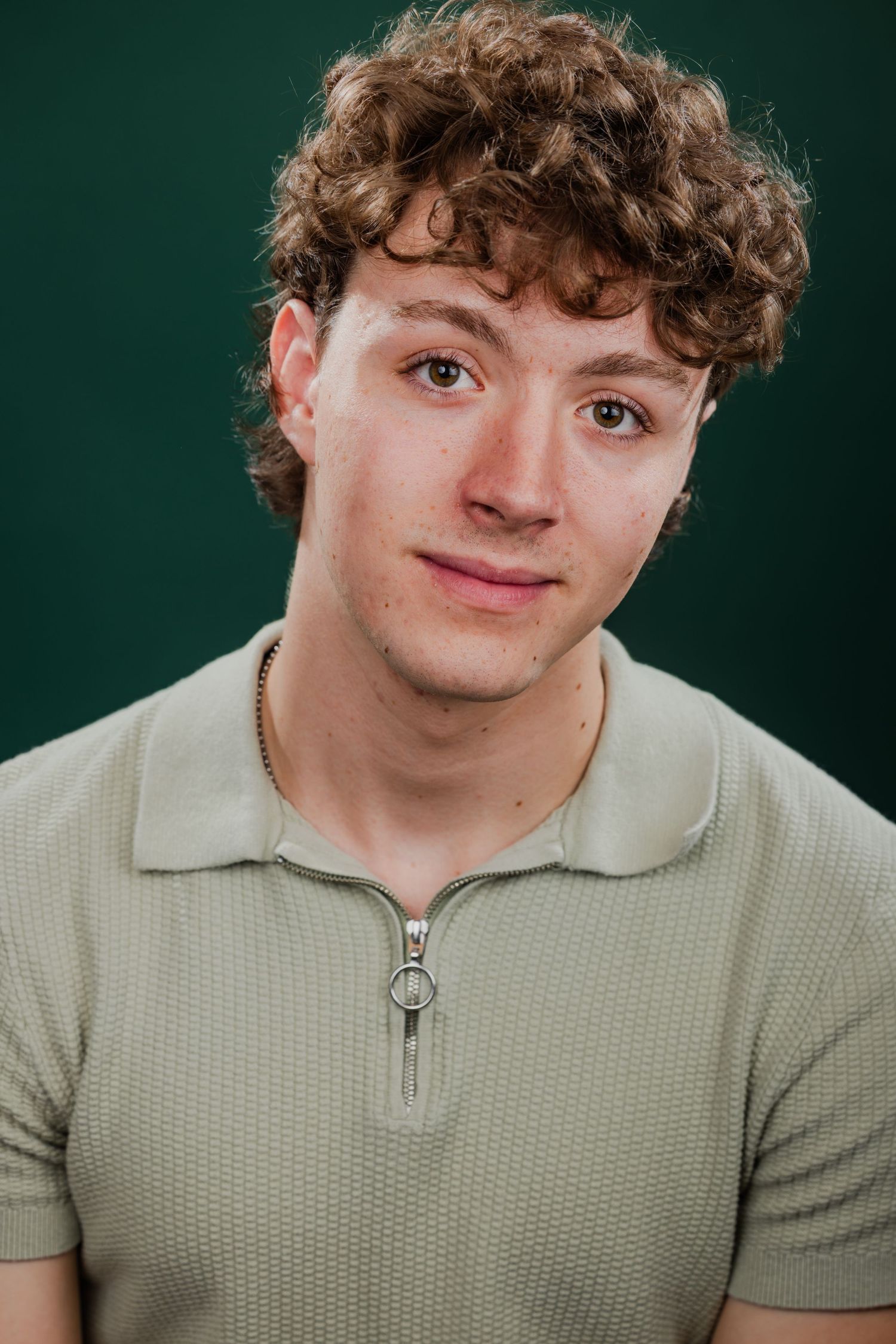 Young man with curly hair wearing a sage green zip polo shirt against a green background.