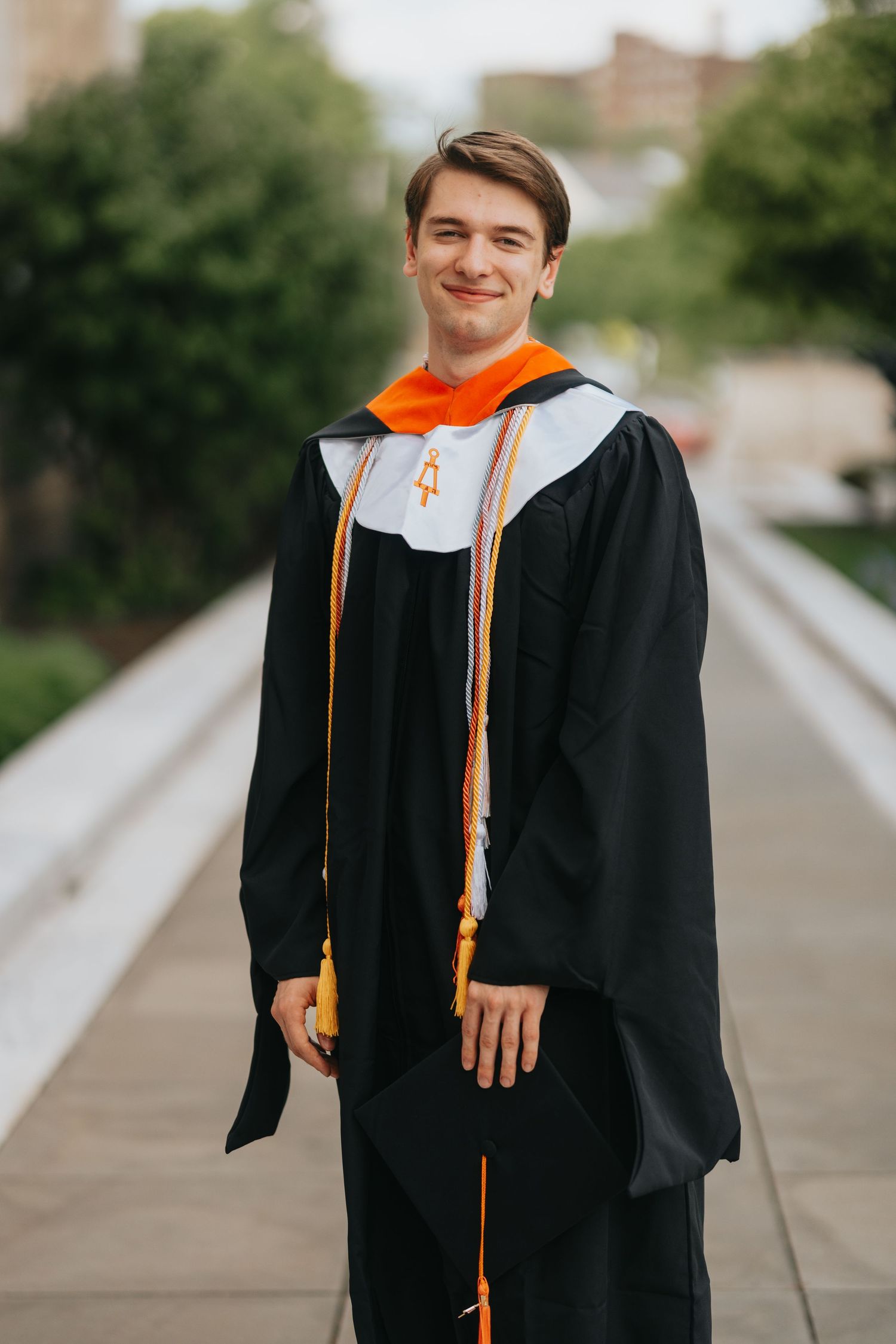 Young man smiling in black graduation gown with orange hood holding diploma outdoors.