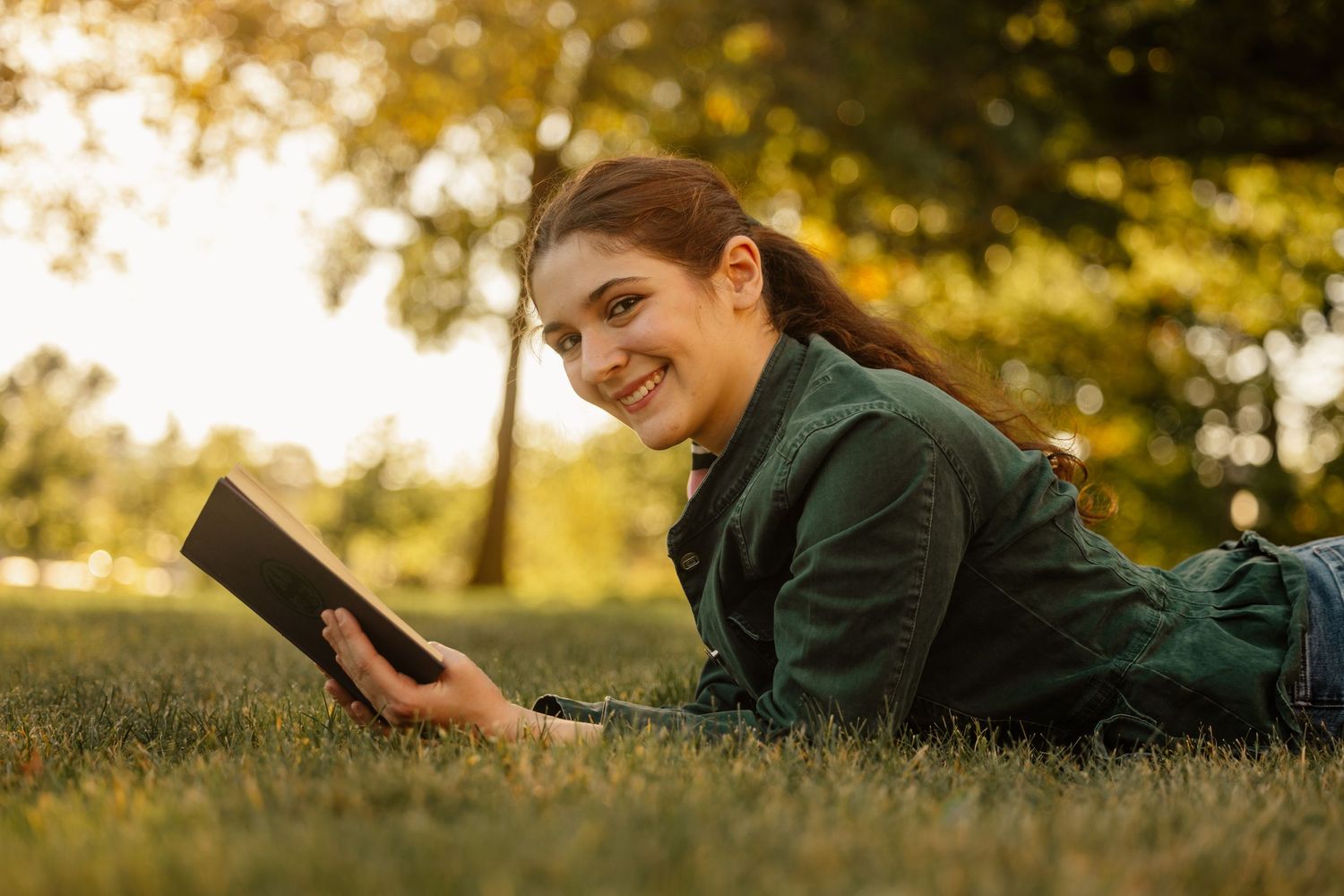 Smiling young woman lying on grass in park reading a book at sunset.