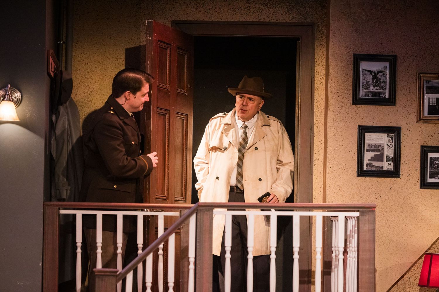 Two men in formal attire converse on a theatrical stage set designed as a home entrance with stairs.