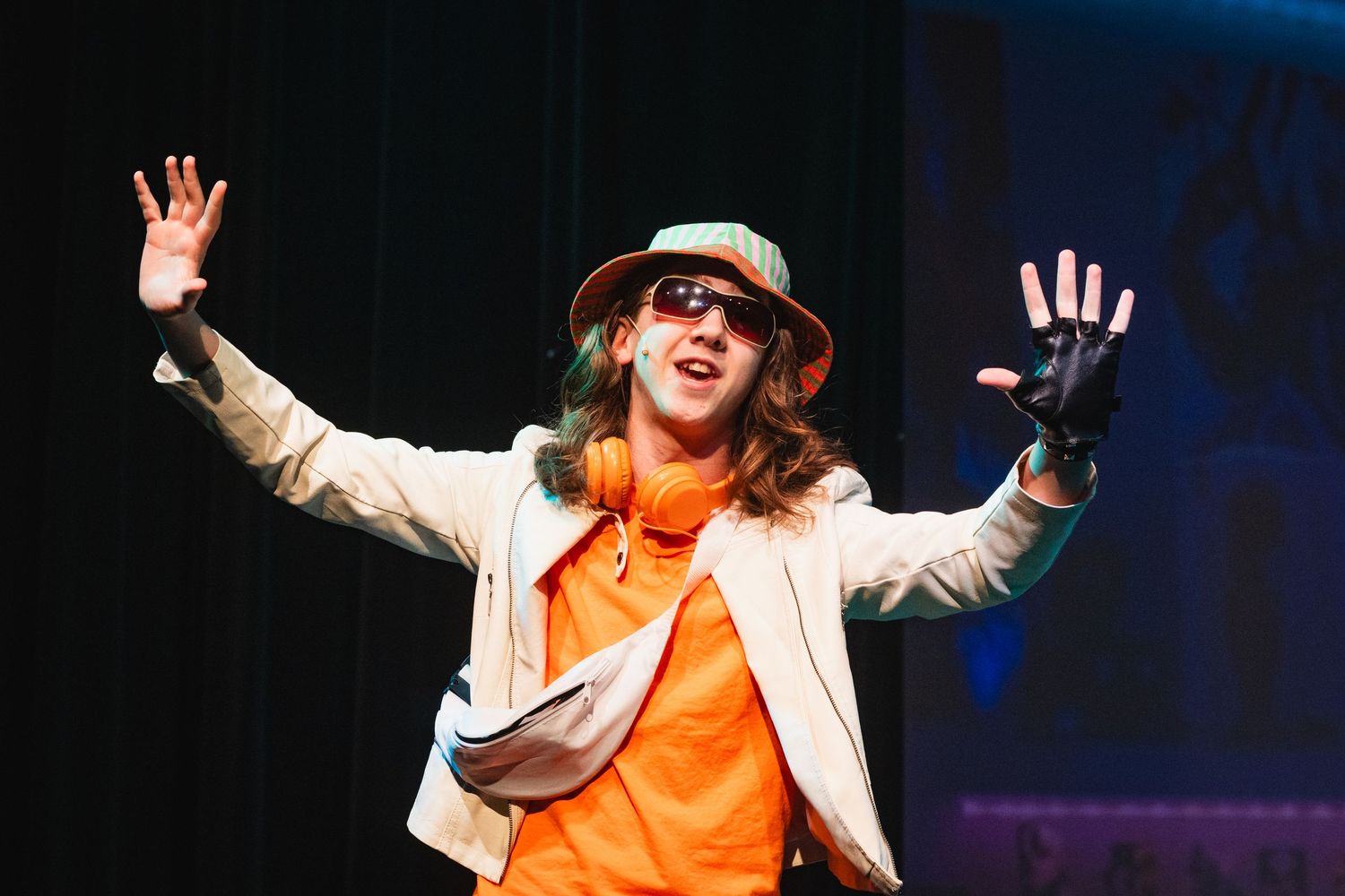 A performer in an orange shirt, bucket hat, and sunglasses stands with arms raised wide on stage.