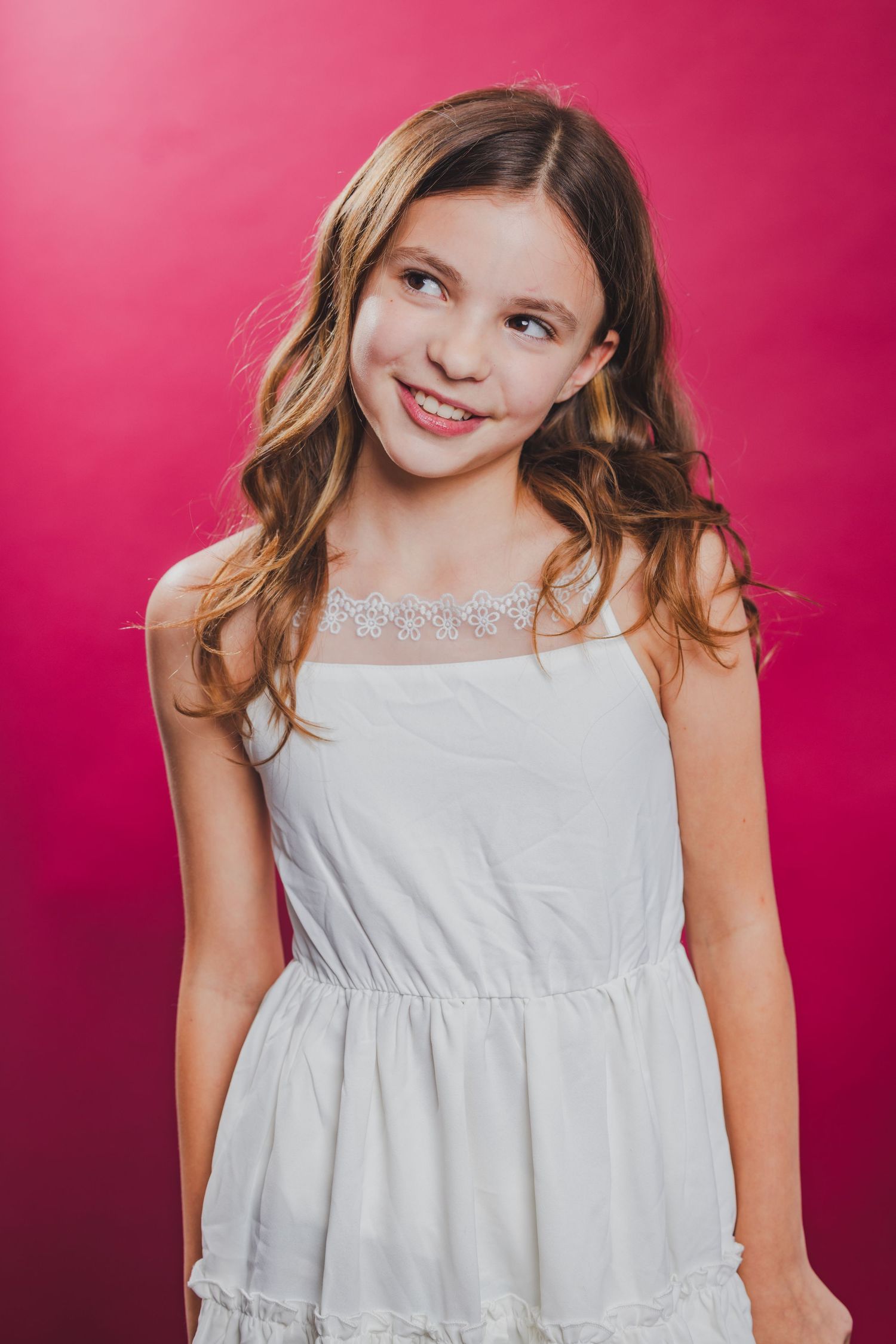 Young girl with wavy brown hair in a white sundress smiling against a pink background.