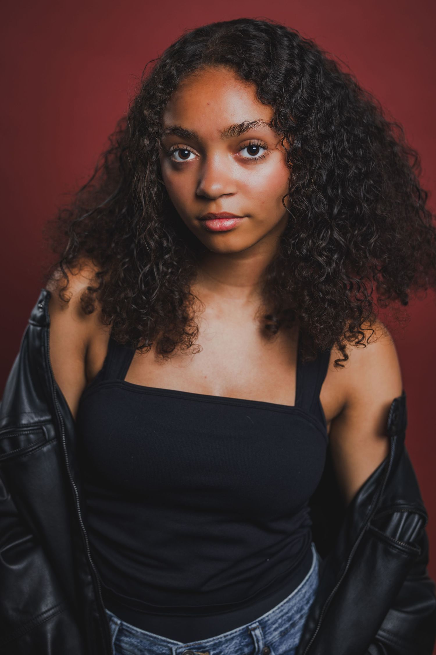 Young Black woman with curly hair wearing black top and leather jacket on red background.