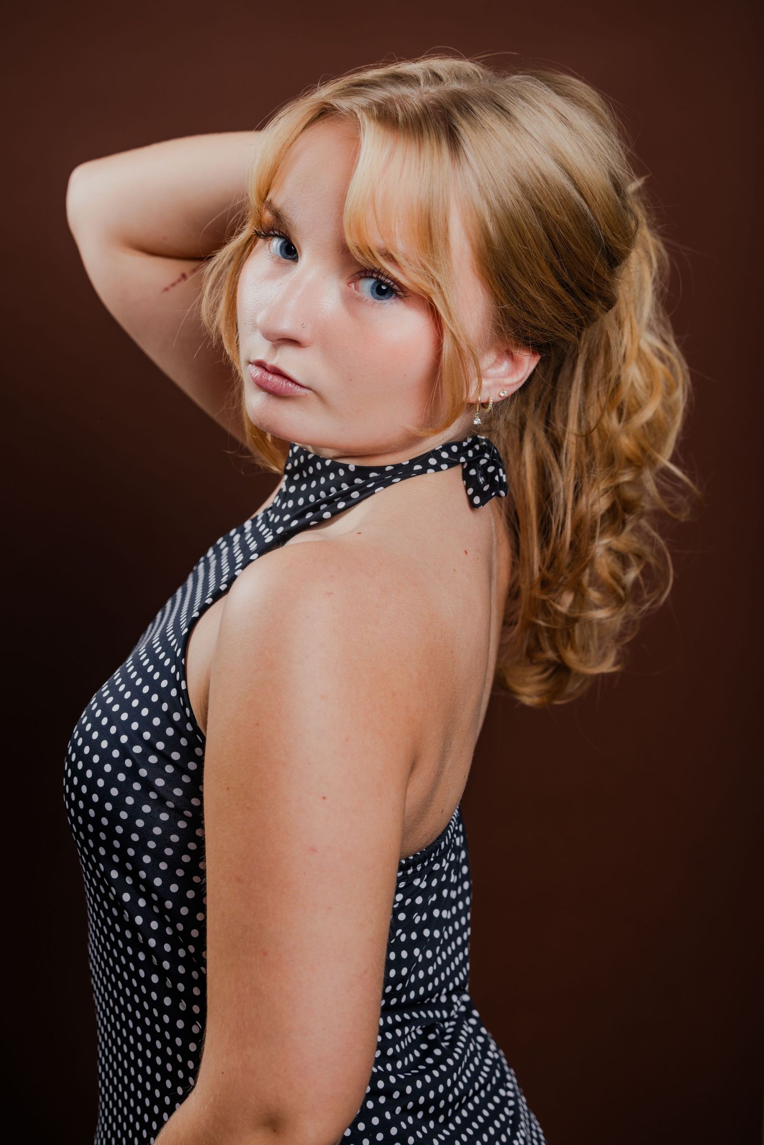 Blonde woman in a black polka dot halter dress posing over her shoulder against a dark brown background.