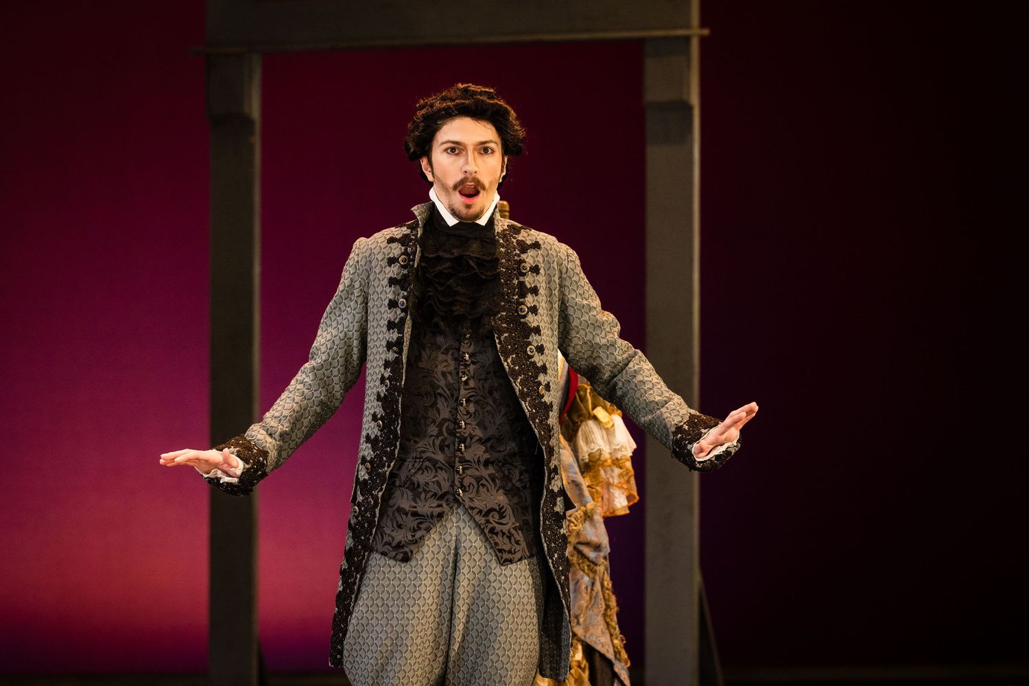 Male opera singer performing on stage in an ornate gray period costume with lace details.